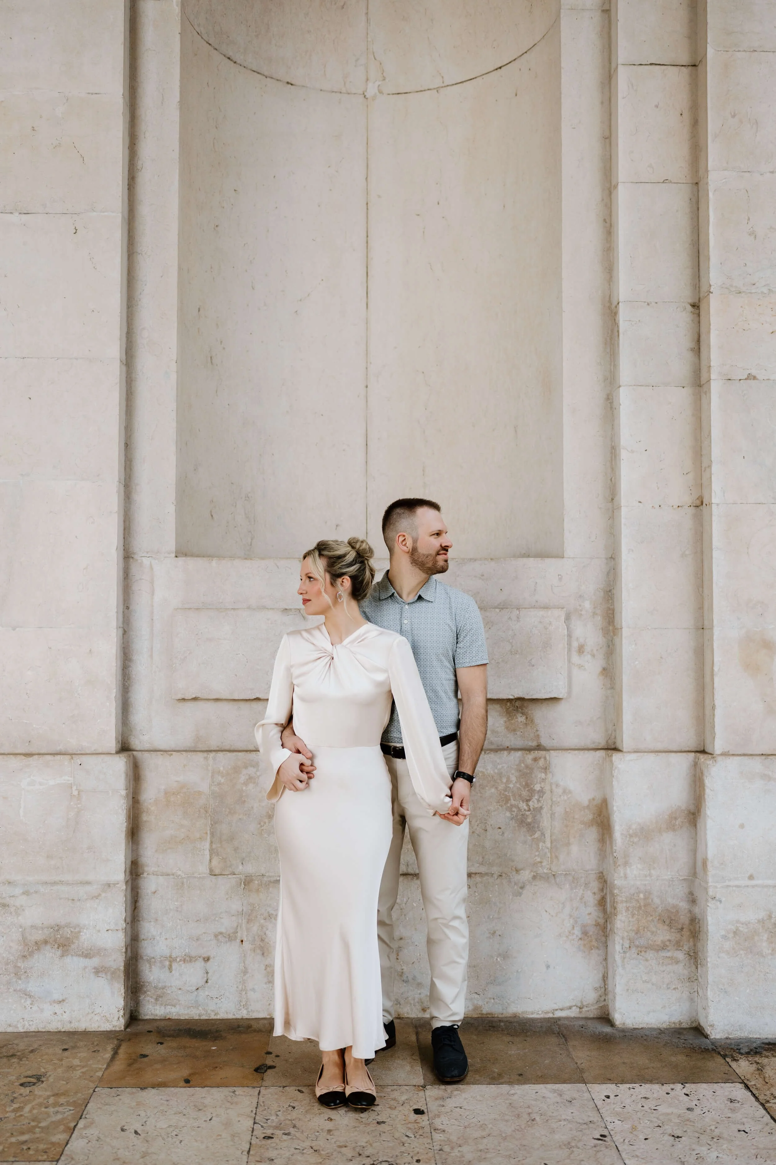 A couple standing against a stone wall, holding hands, with the woman in a white dress and the man in a light blue shirt and cream pants.