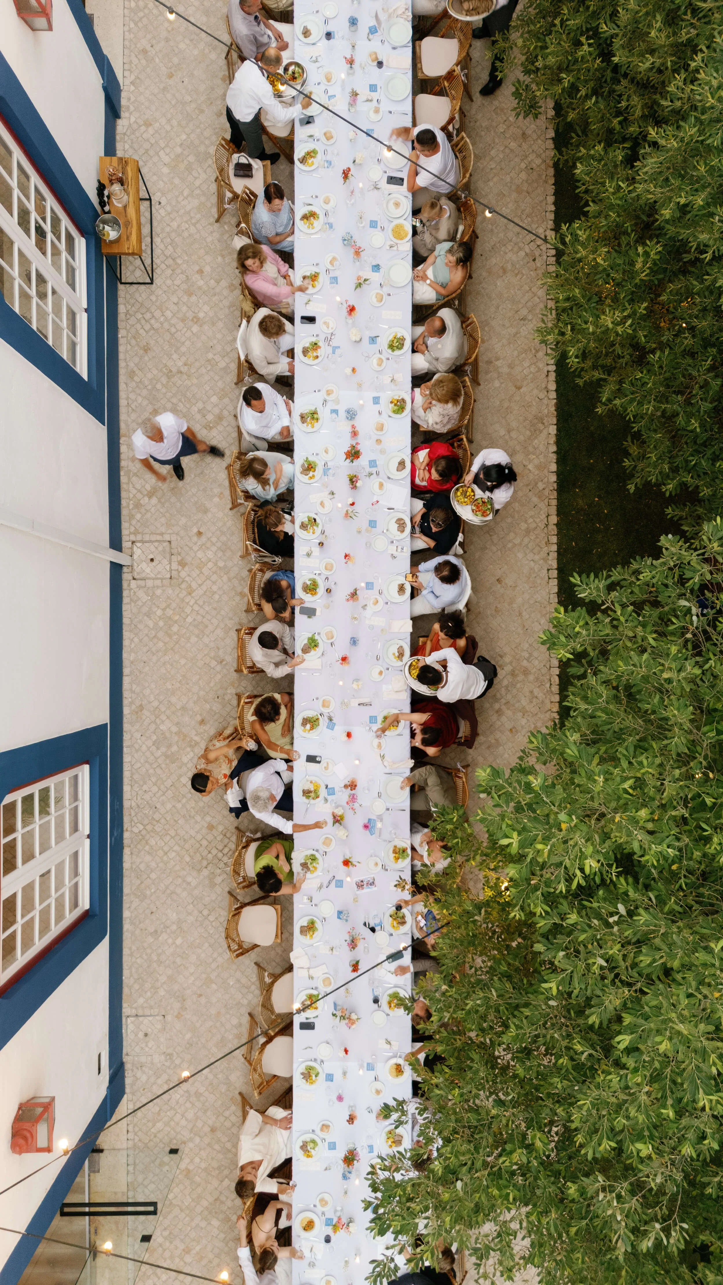 Overhead view of a long outdoor dining table set for a wedding gathering, with people seated around it, on a patio with greenery on one side and a white building with blue trim on the other.