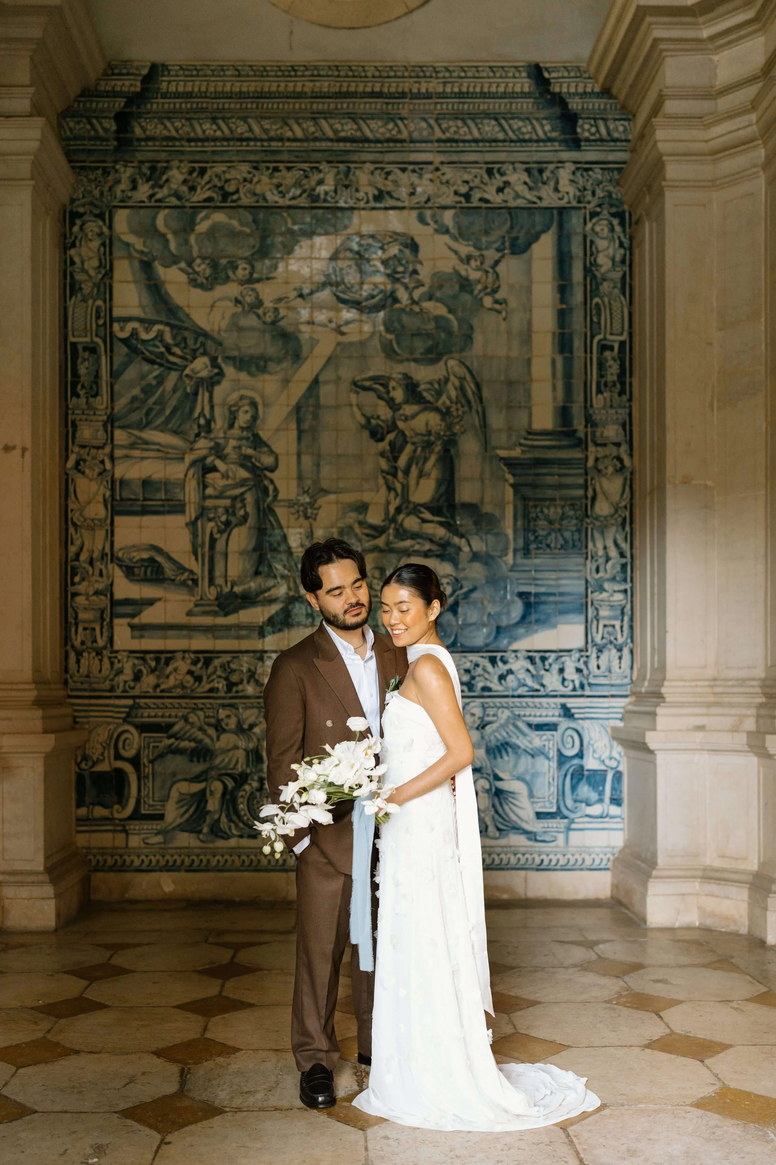 A bride and groom standing close together, smiling, in front of an ornate tiled mural with religious imagery, indoors.