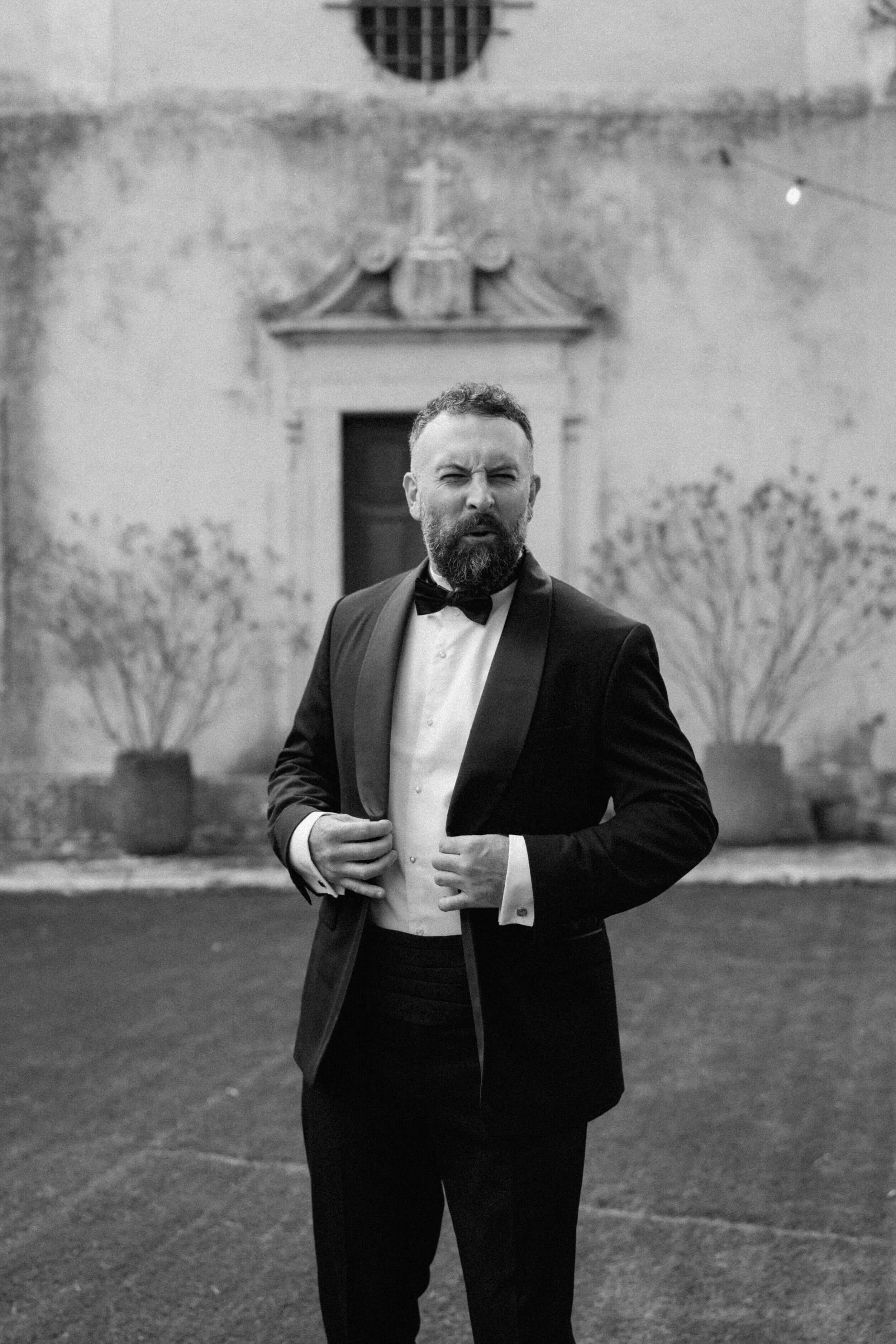 A man in a tuxedo adjusting his jacket outdoors, with a textured wall and potted plants in the background, in black and white.