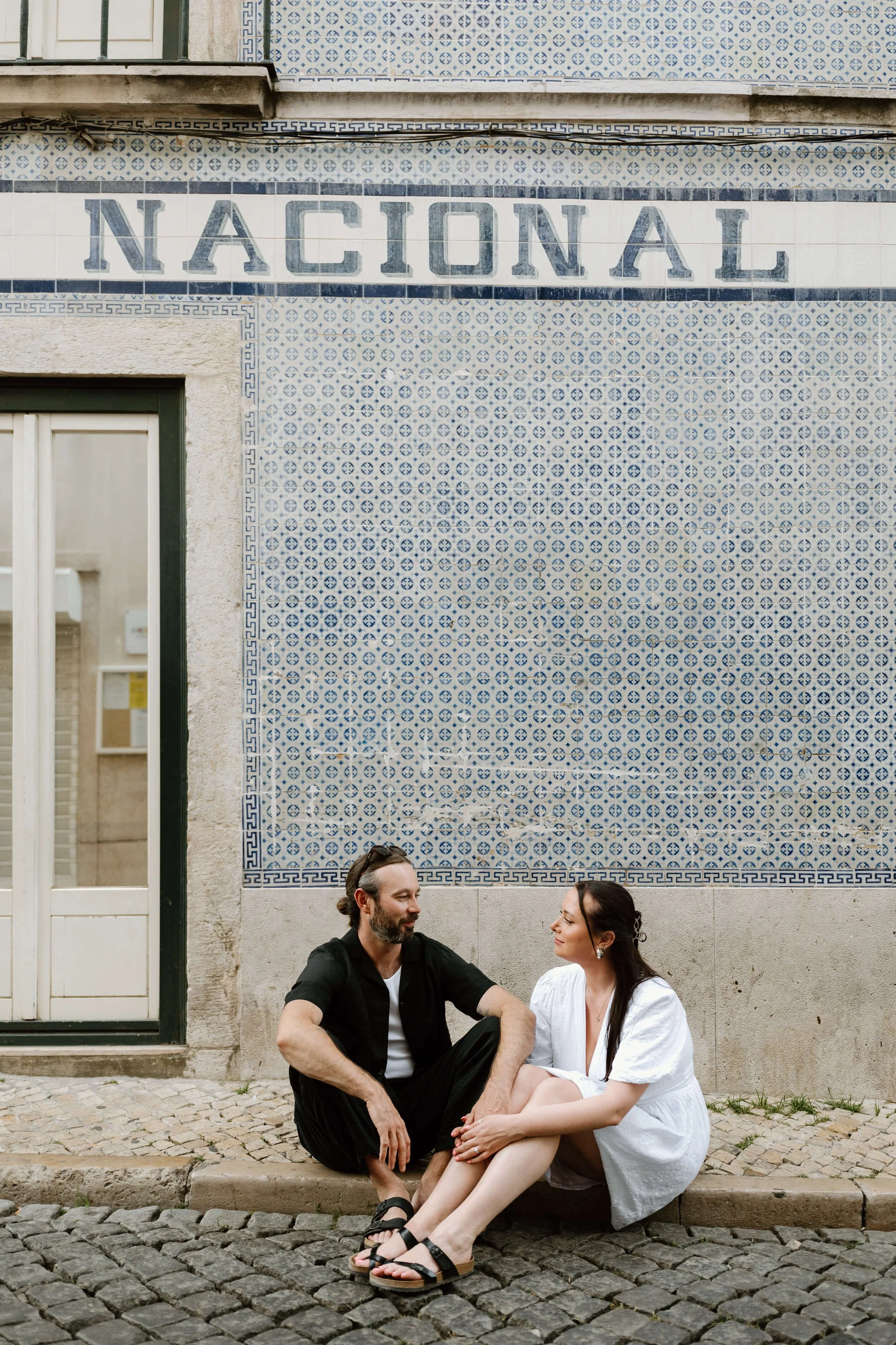 A man and woman sit on cobblestone street in front of a tiled wall with the word 'NACIONAL' painted on it, engaging in conversation.