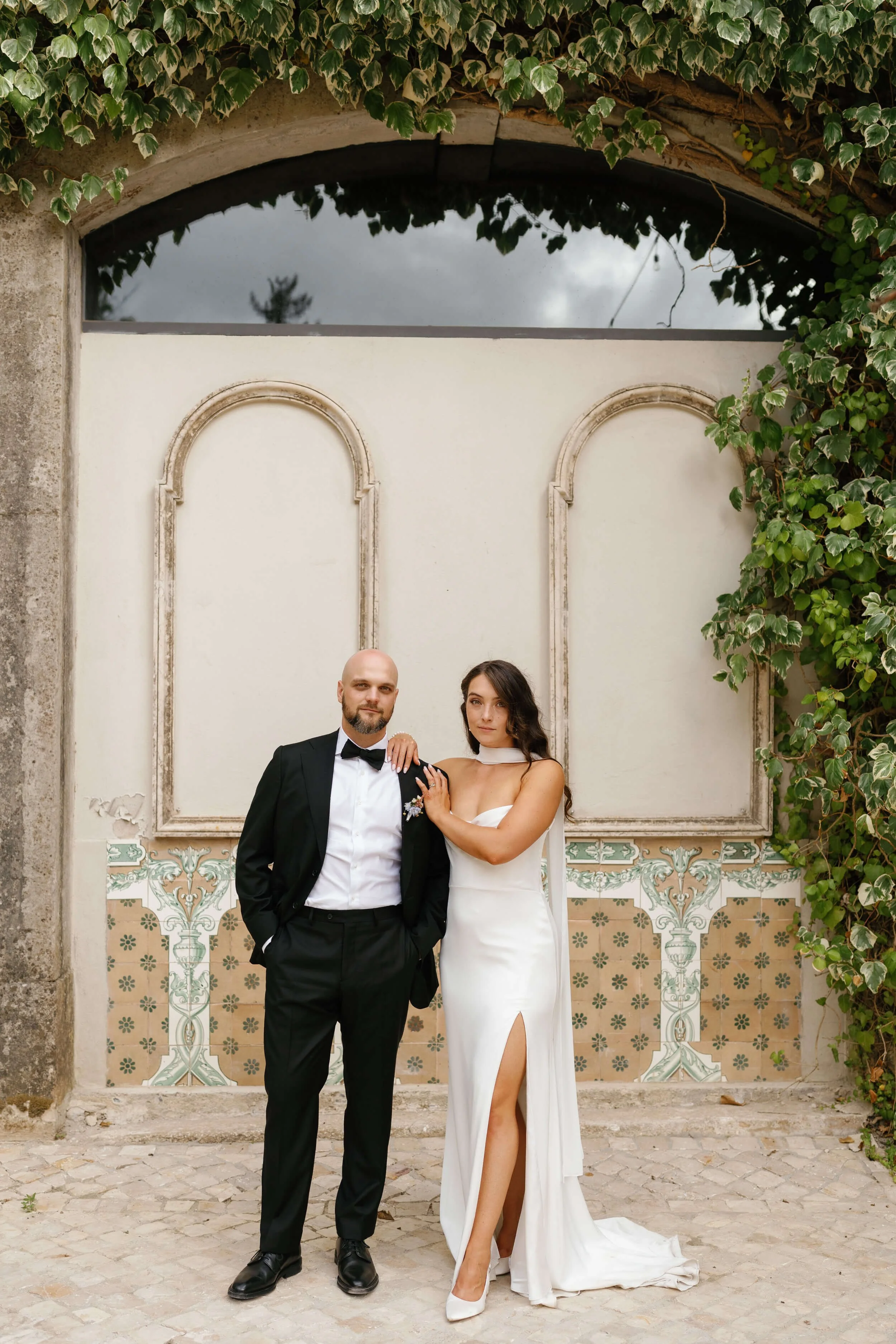 A couple in wedding attire standing outdoors, with the man in a black tuxedo and the woman in a white gown with a thigh-high slit, against a wall with decorative tiles and greenery.