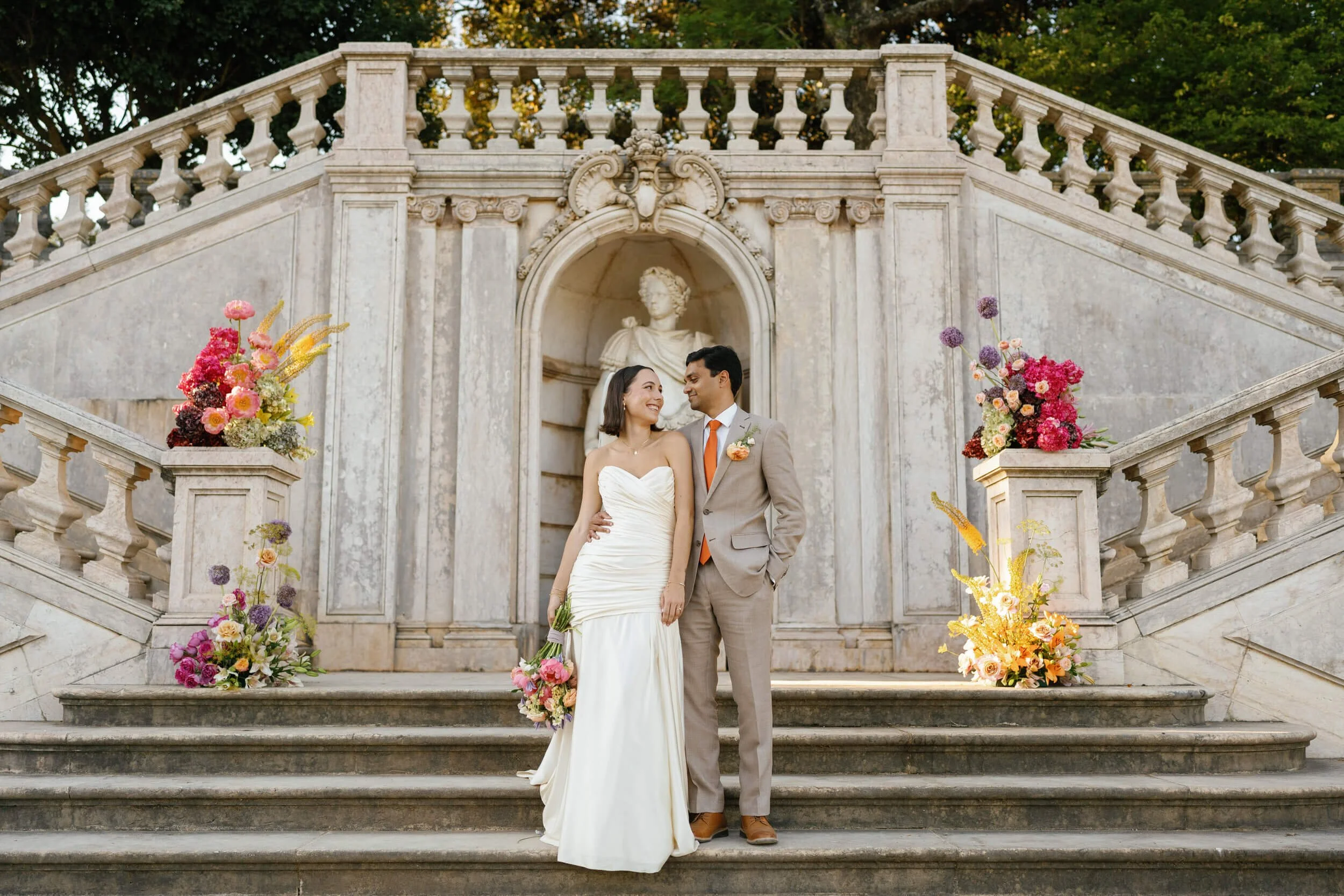 Brazilian bride and Indian groom posing in the iconic Vandelli Botanical Gardens staircase, framed by Greenpick'd florals full of pink and orange tones