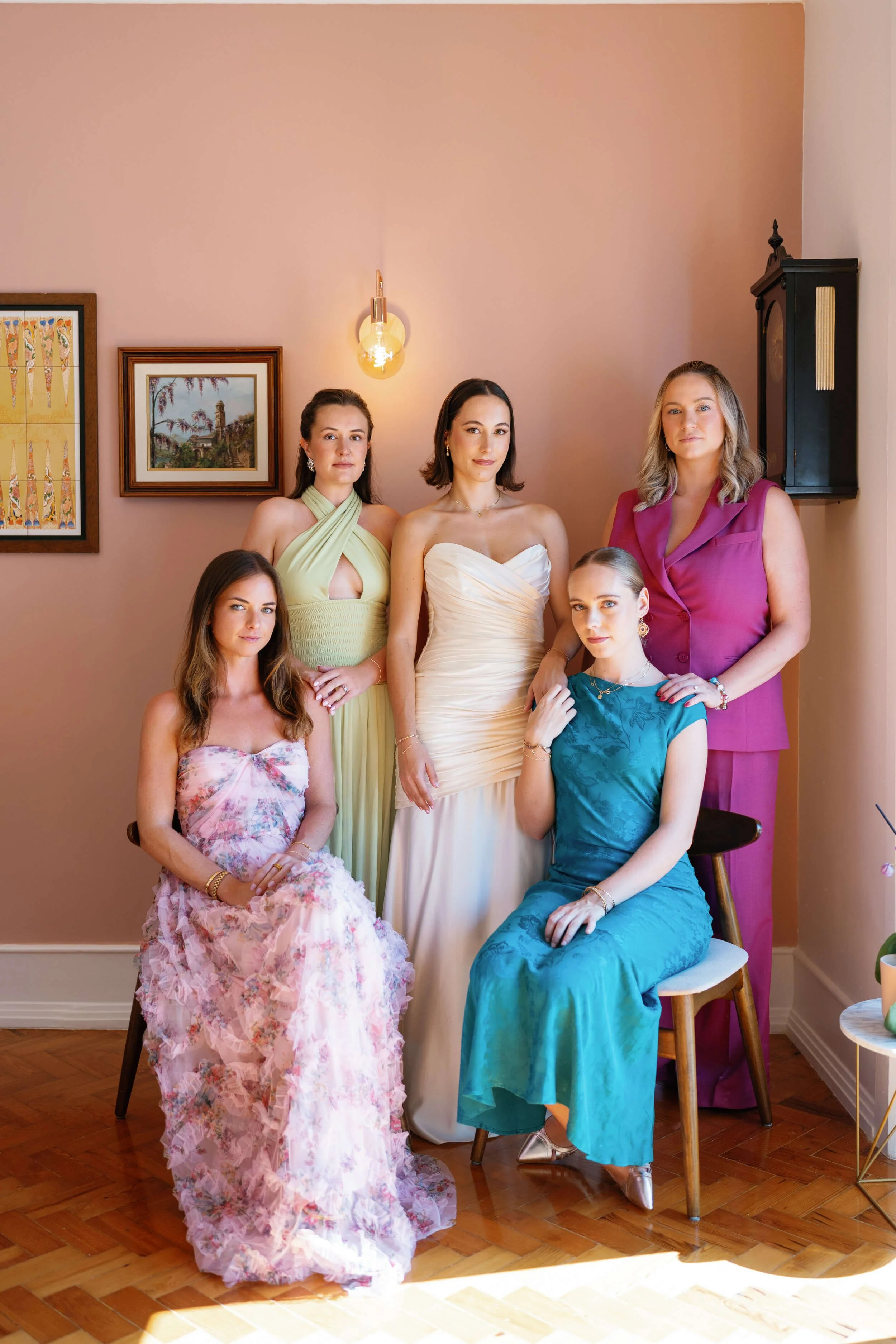 Six women dressed in colorful formal dresses and outfits, posing together in a room with pink walls and artwork.