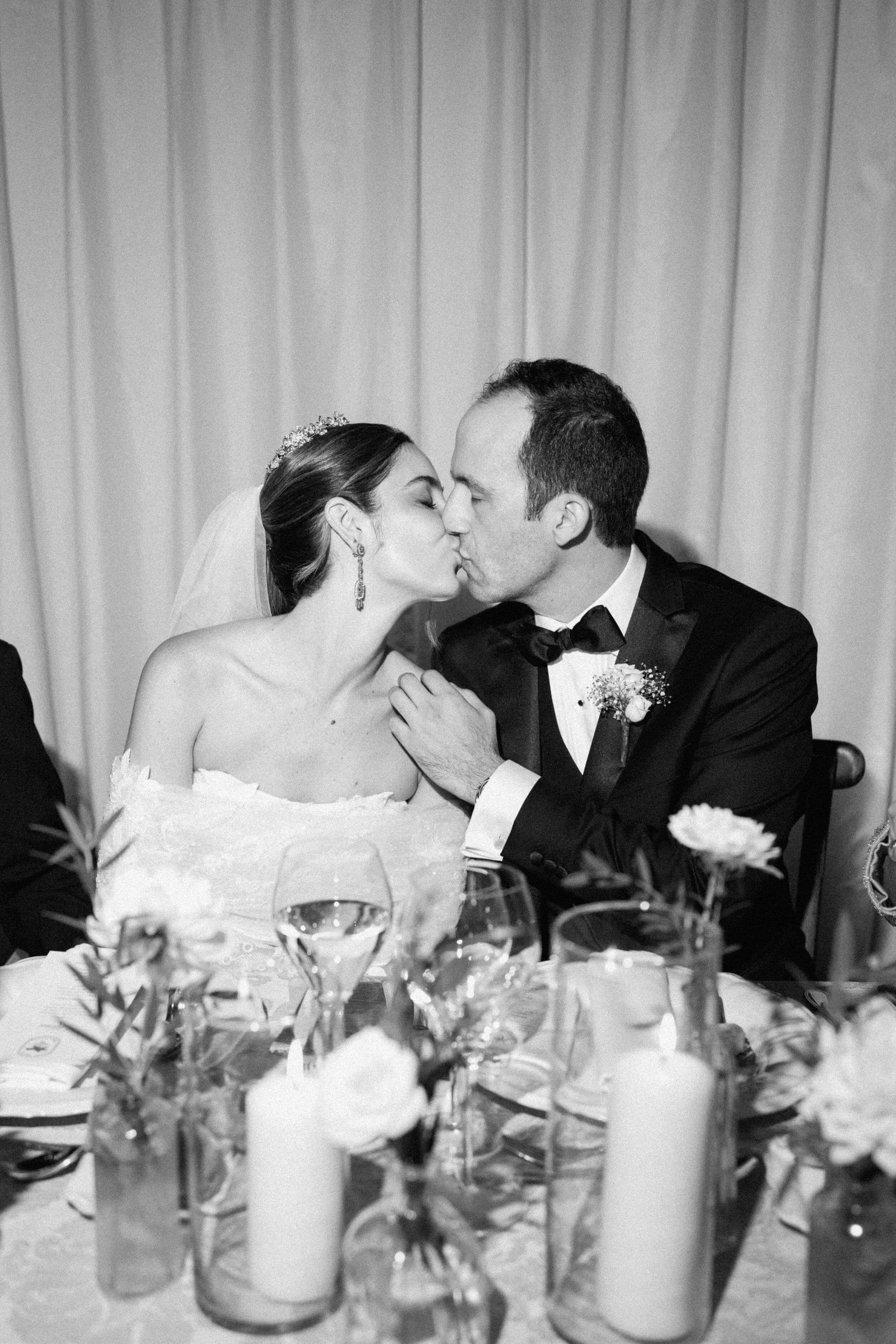 Black and white photo of a bride and groom sharing a kiss at their wedding reception, seated at a decorated table with flowers and candles.