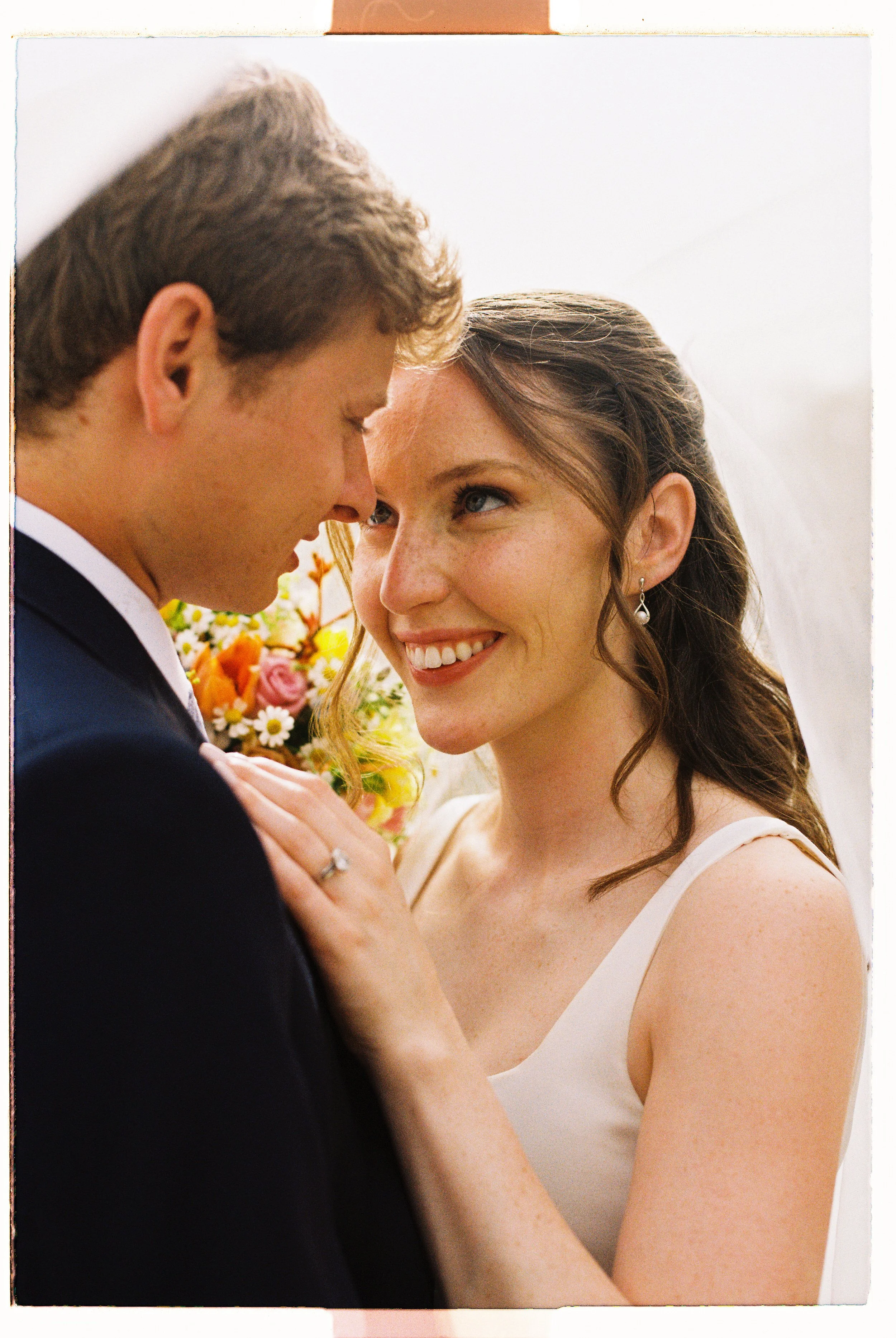A close-up of a happy couple at their wedding, with the bride smiling and the groom with their foreheads touching, holding hands in front of a floral bouquet with a white background.
