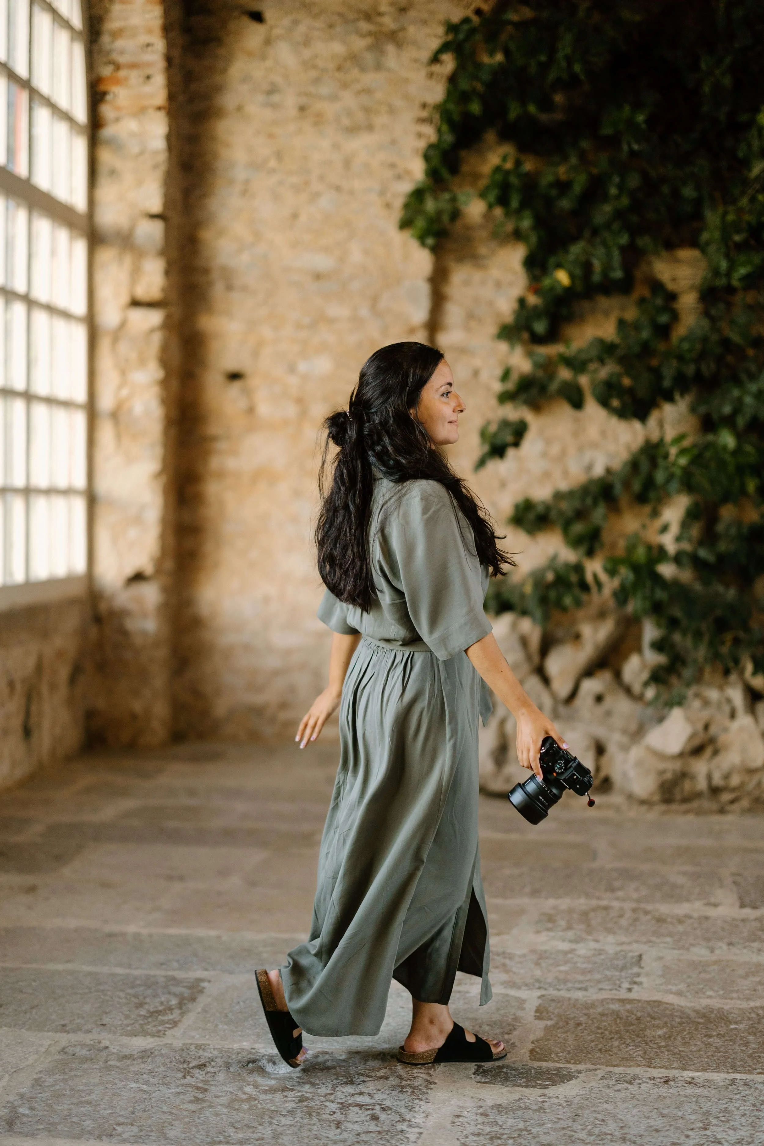A woman with long dark hair walking indoors, holding a camera, in front of a rustic stone wall with a large window and green foliage.