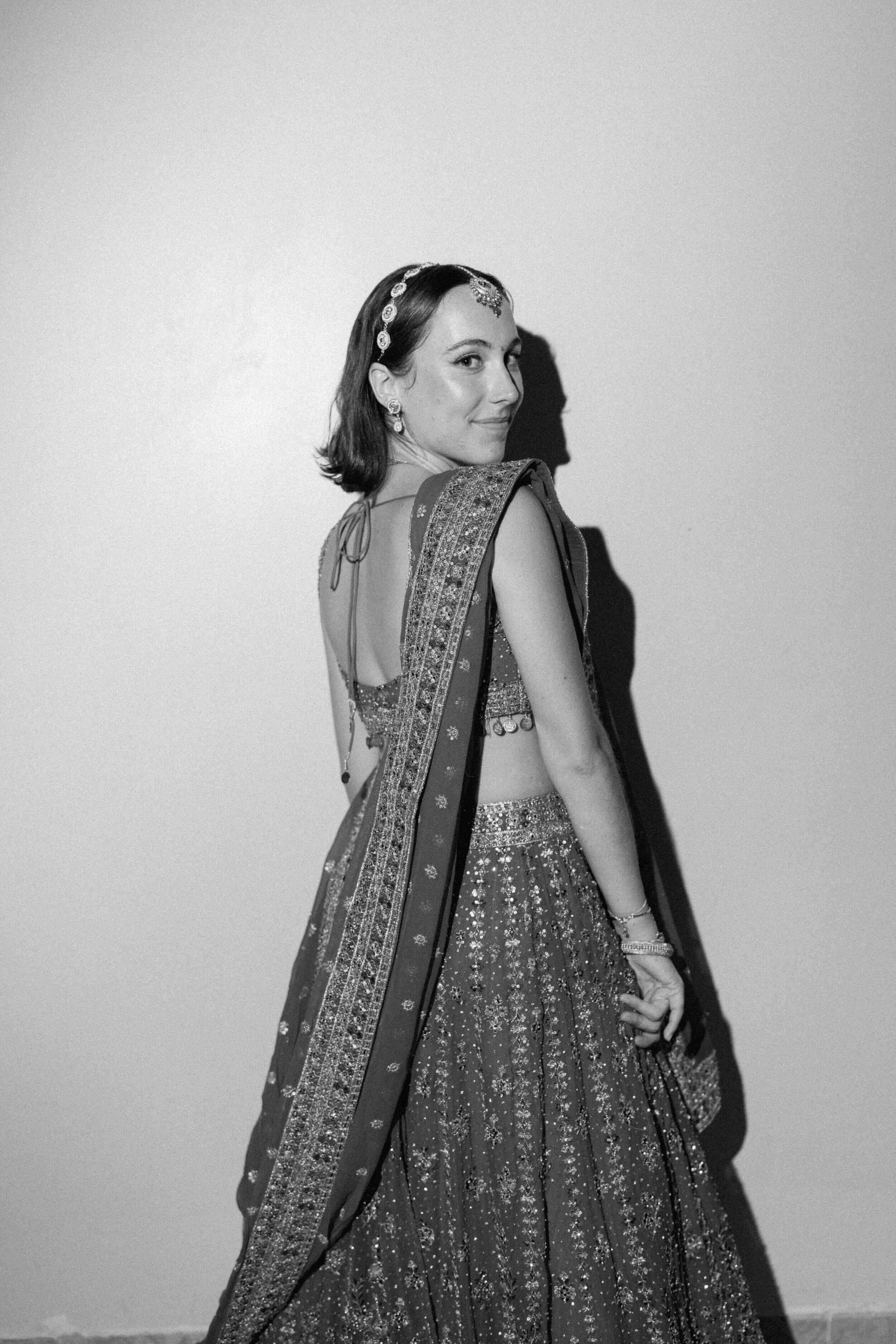 A woman dressed in traditional Indian attire, including a heavily embroidered saree or lehenga, with jewelry and a headpiece, standing against a plain wall.