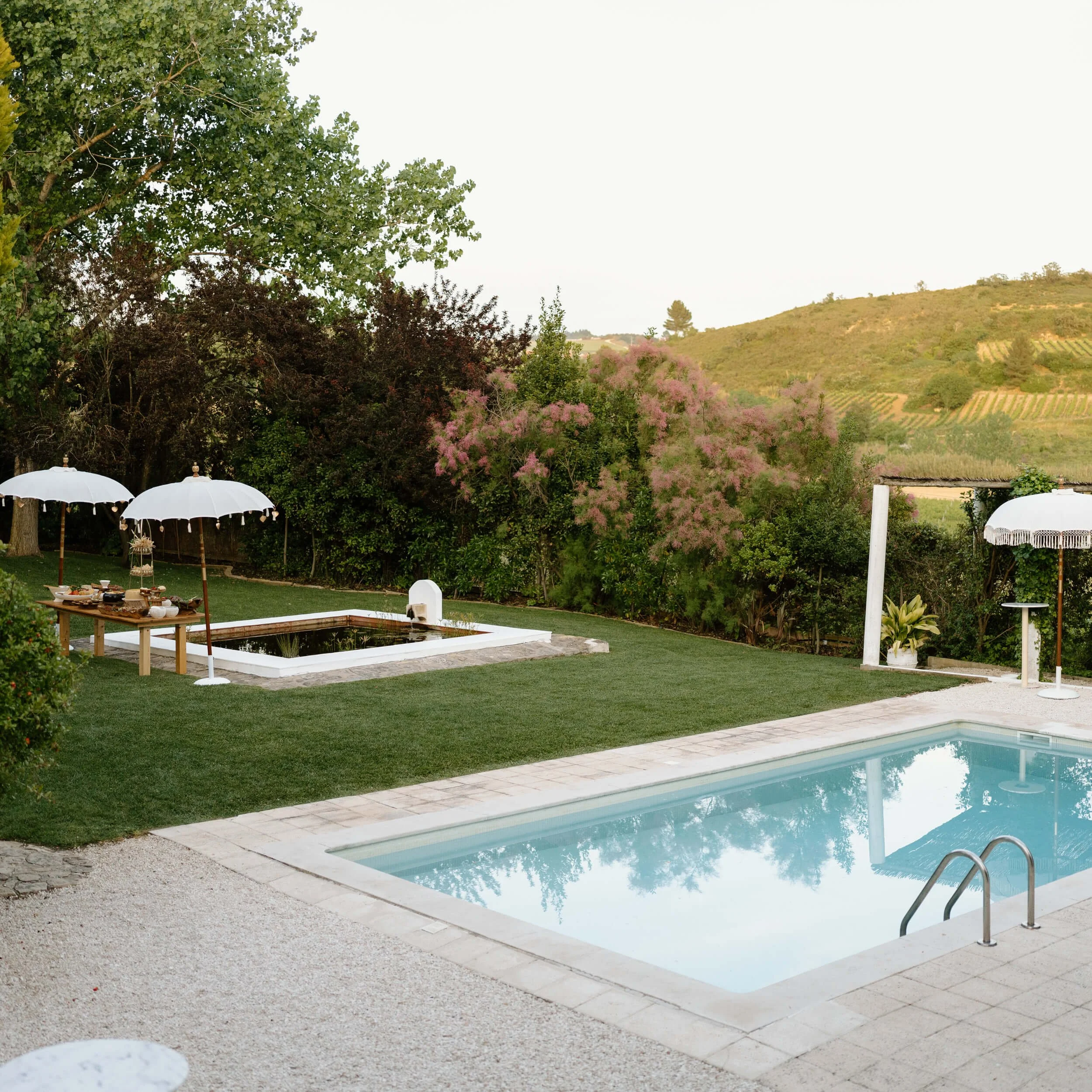 A backyard pool with a ladder, surrounded by a stone deck and green grass, with white umbrellas, a picnic table, and trees in the background.