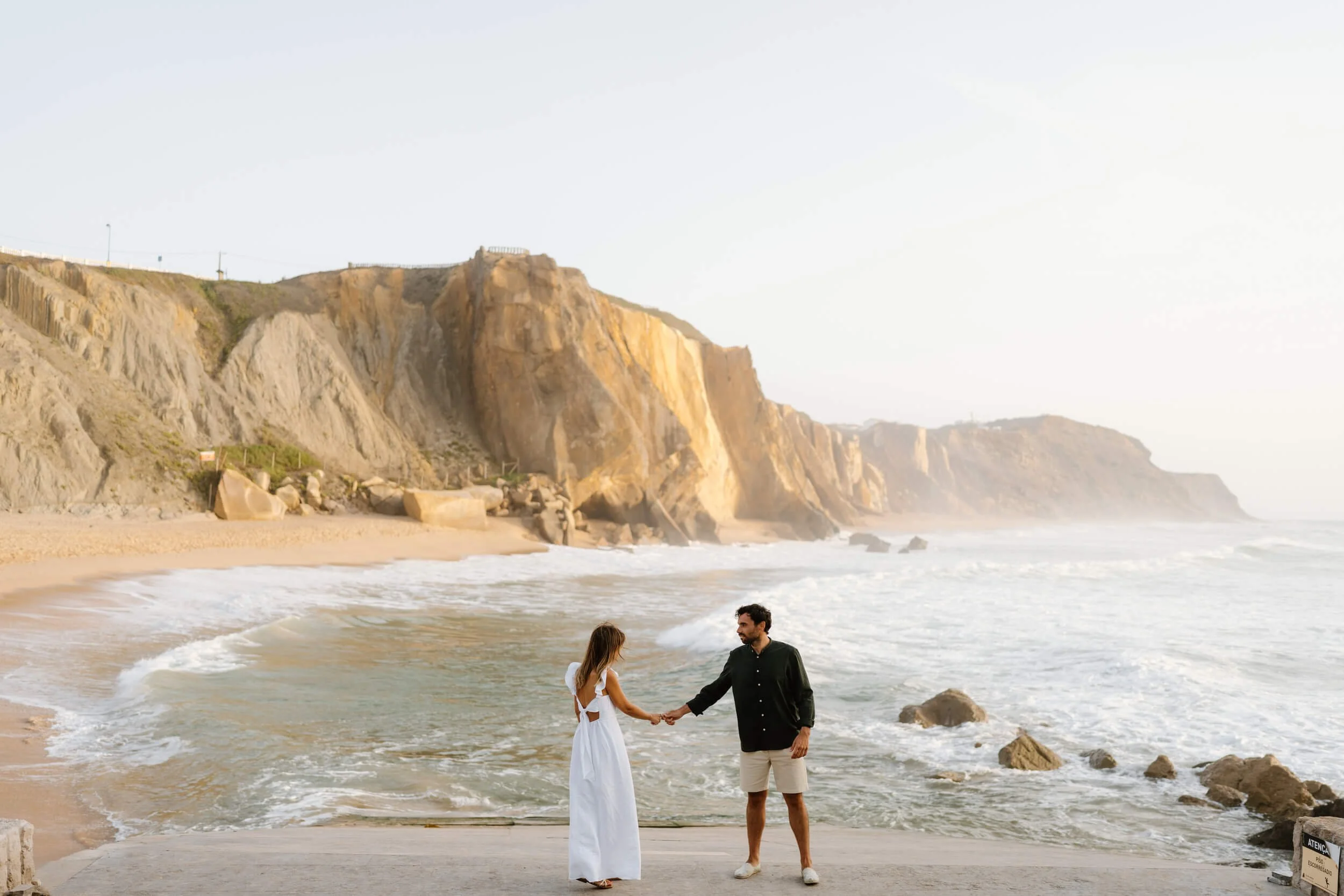 A man and woman holding hands on a beach with waves and cliffs in the background during sunset.