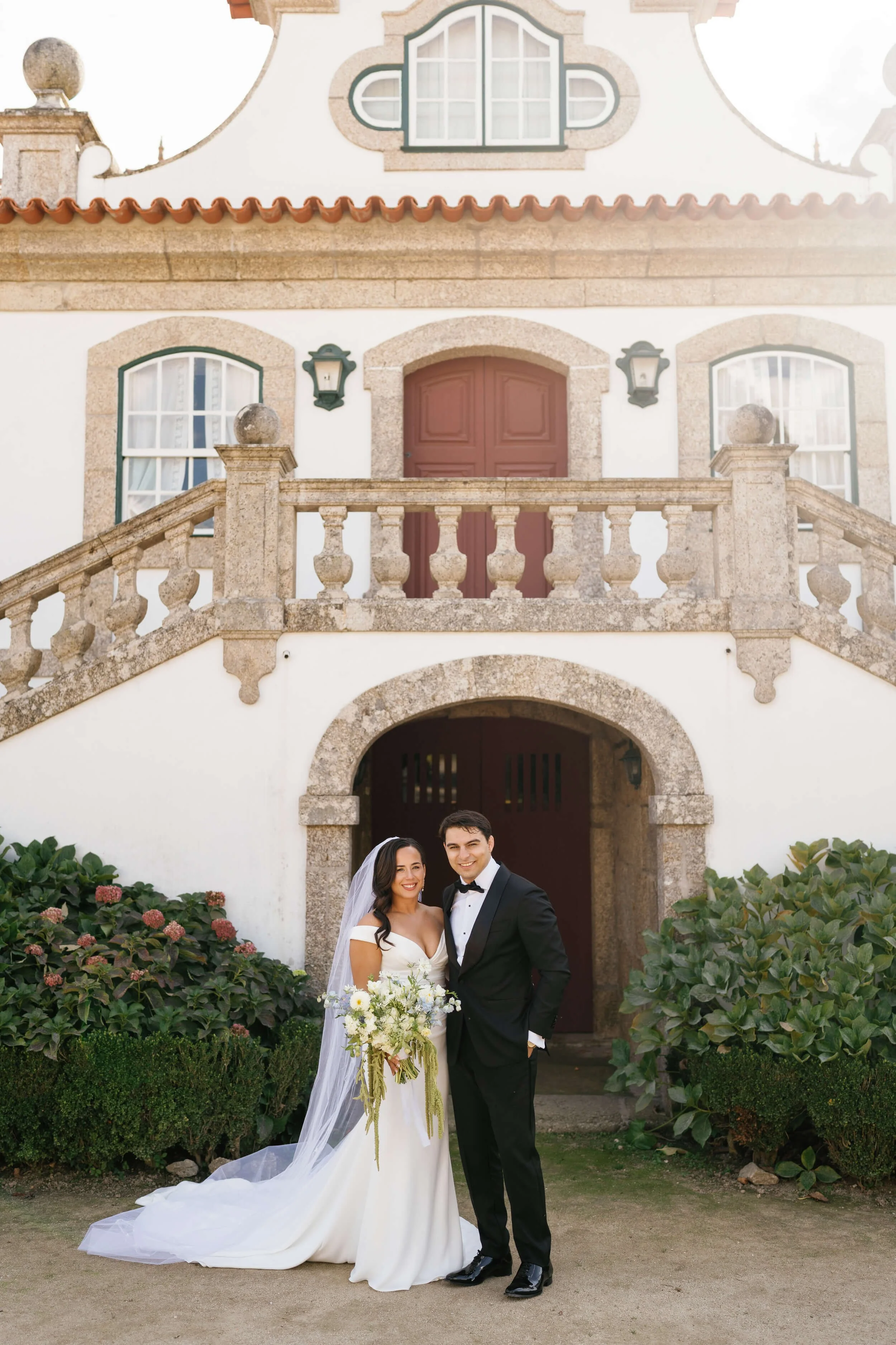 Bride and Groom standing in front of Casa Vila Verde, a 13th century manor in the north of Portugal. Posing and smiling to the camera, while the bride holds her bouquet with white flowers and green amaranthus.