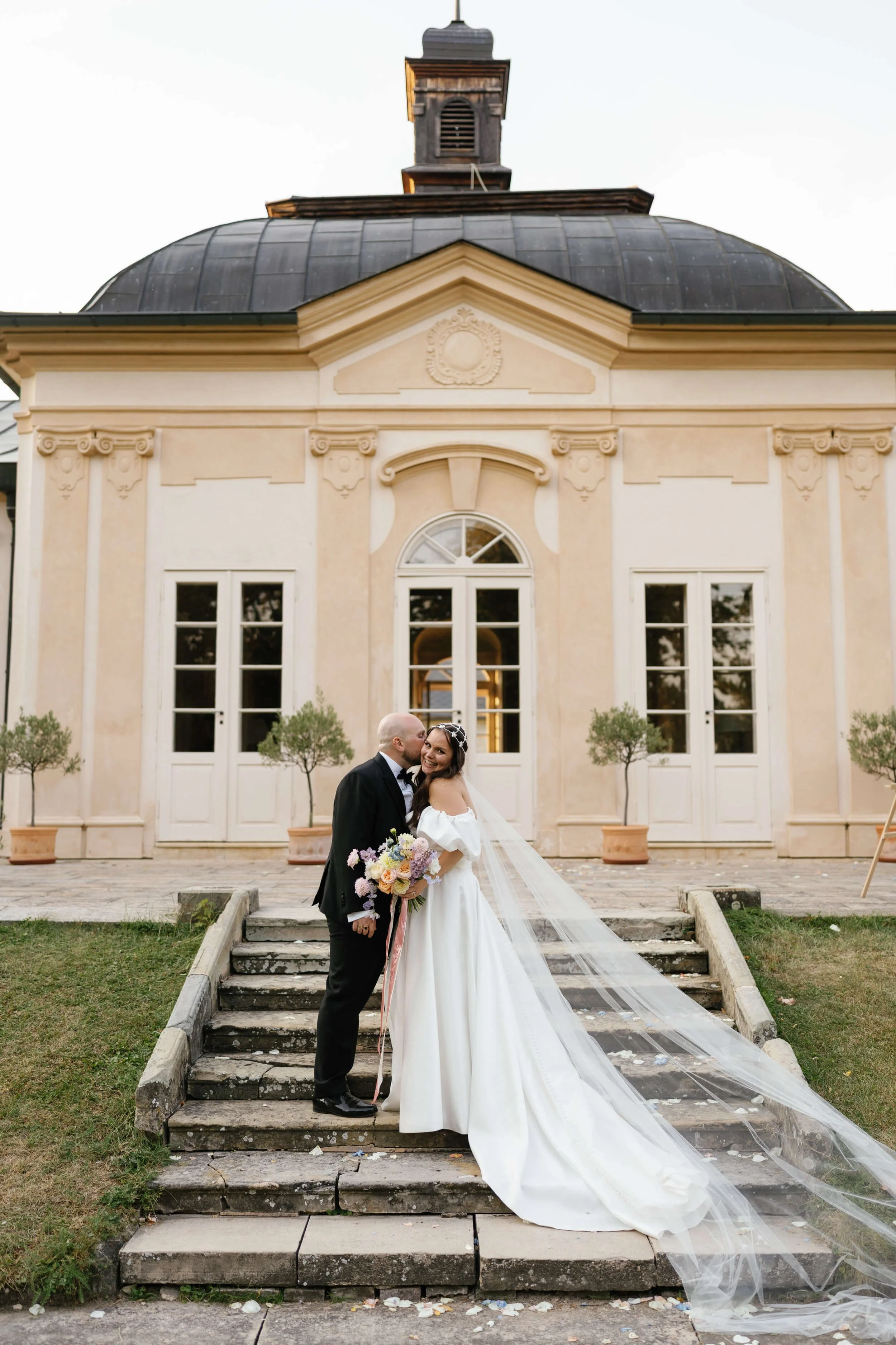 A bride and groom stand on stairs outside a historic church, with the bride holding a bouquet of flowers, smiling, and the groom leaning in to kiss her.