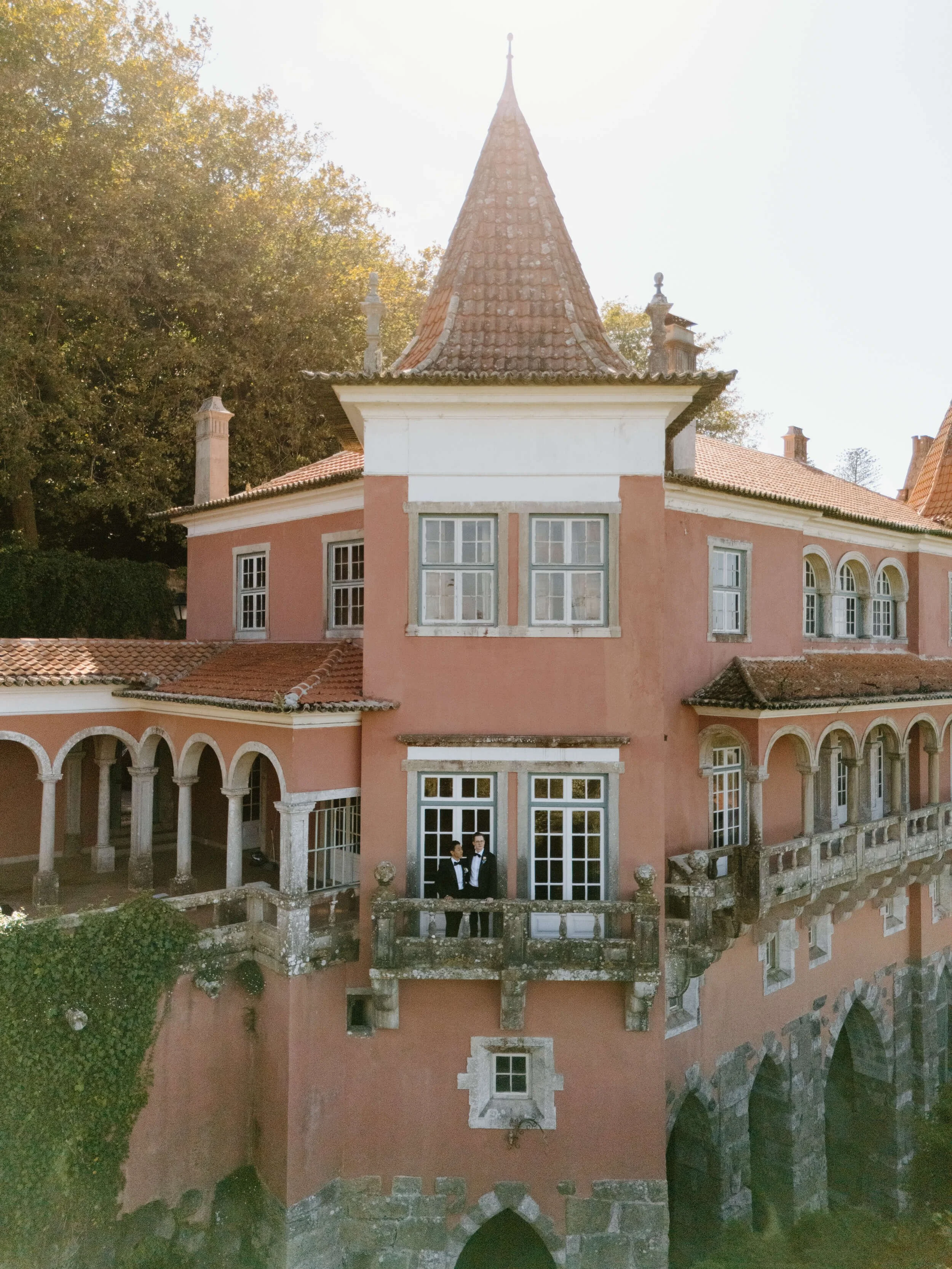 A large pink castle with a turret and balconies, surrounded by trees, with two men in tuxedos standing on one of the balconies.