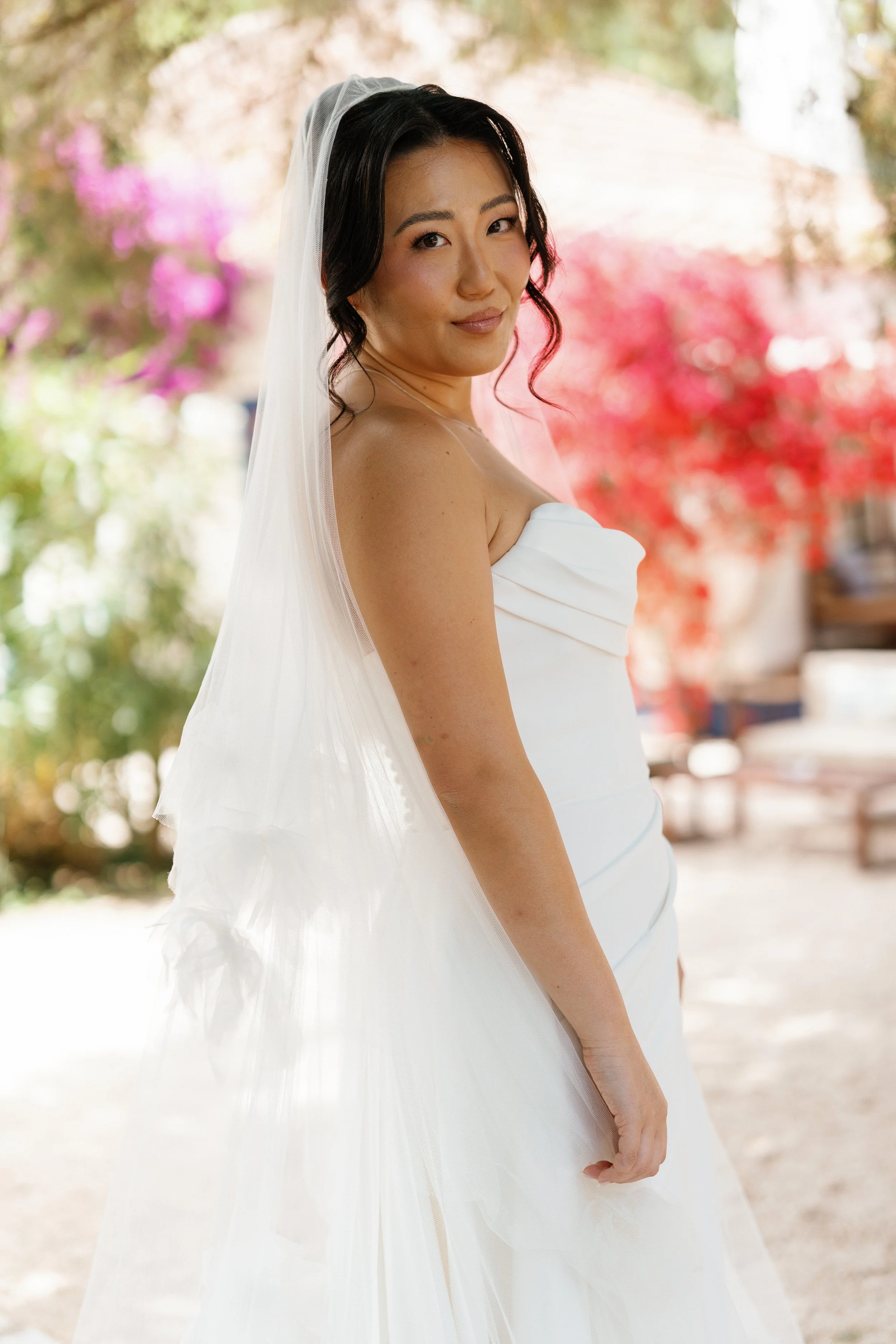 A woman in a white strapless dress and veil, standing outdoors with pink and purple flowers in the background.