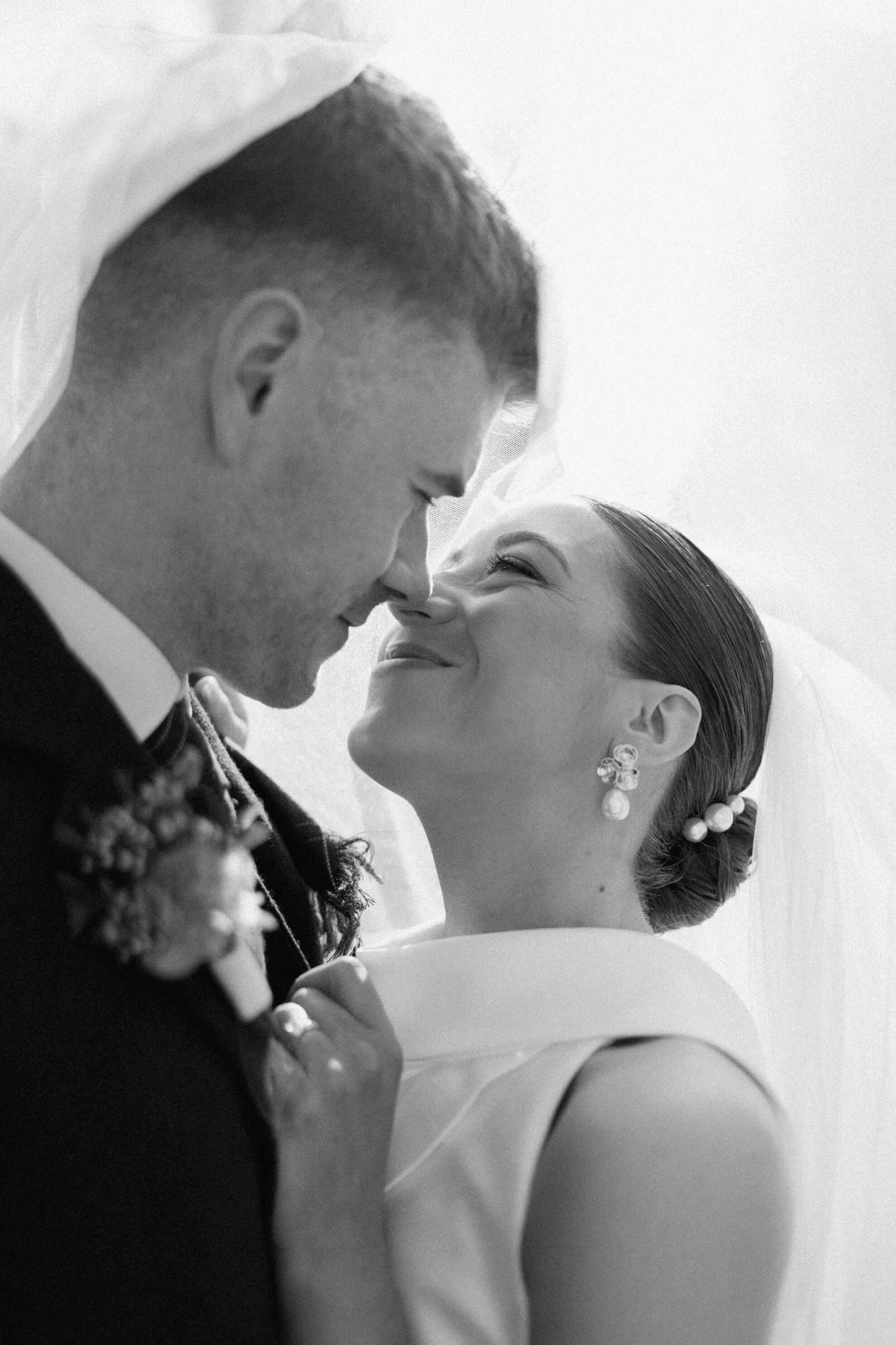Black and white photo of a bride and groom touching noses, smiling with eyes closed. The bride has her hair in an elegant updo with pearl earrings and a veil, wearing a sleeveless dress. The groom wears a tuxedo with a boutonnière, leaning in closely
