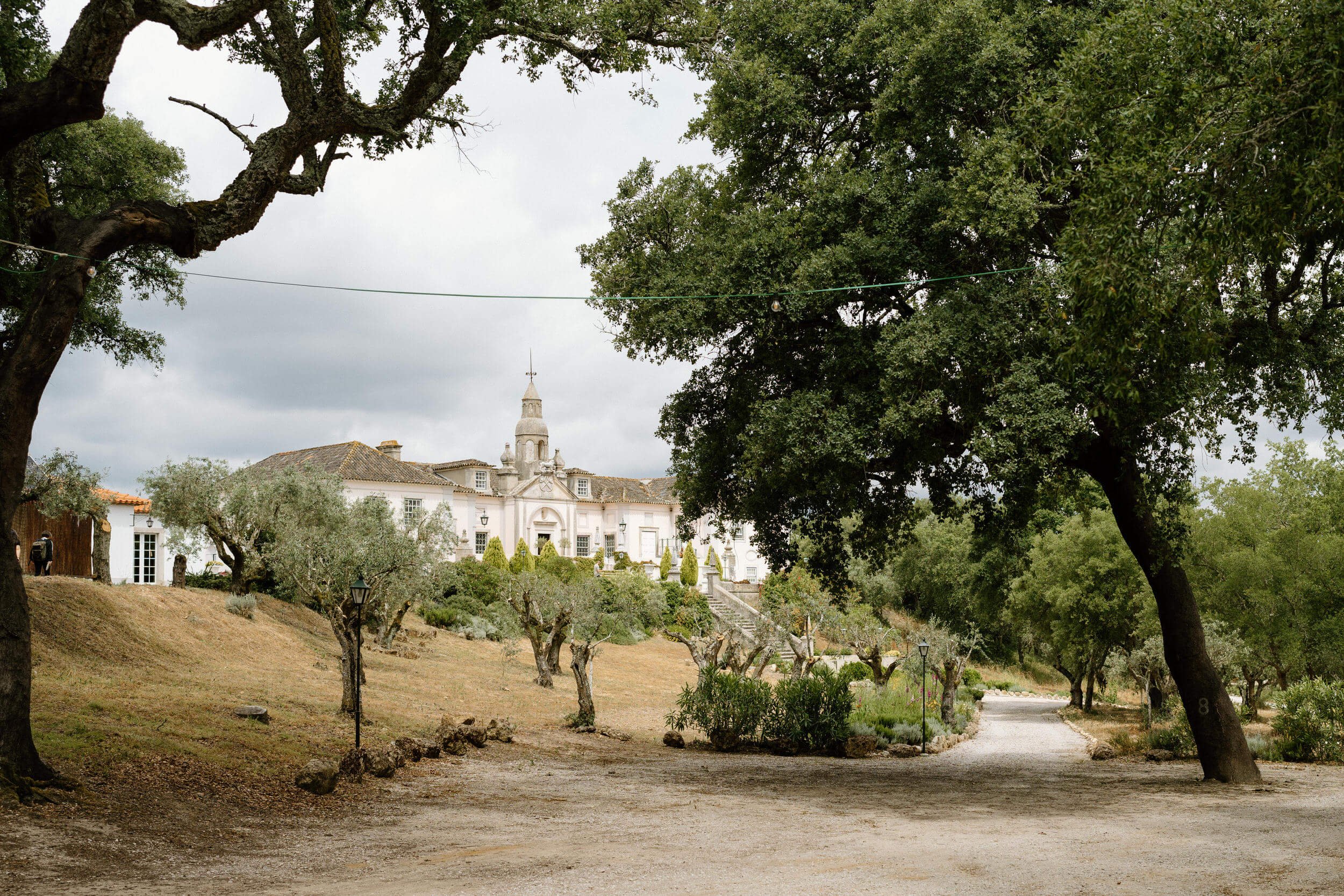 A scenic view of a white mansion with a tower, surrounded by trees and a dirt pathway, under a cloudy sky.
