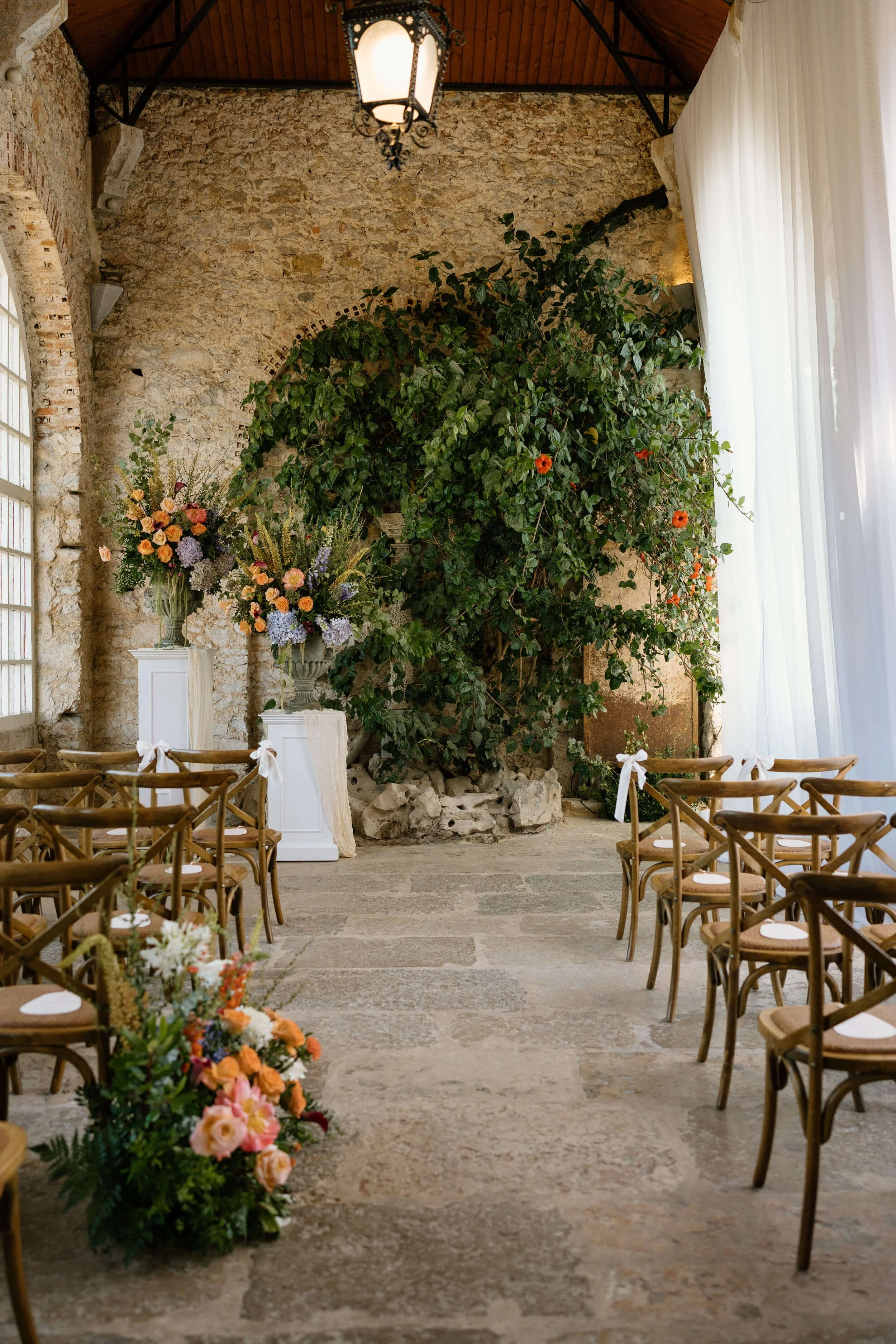 Interior of a rustic wedding ceremony space with wooden chairs, large floral arrangements on white pedestals, a large green leafy plant, and a stone wall with large windows and white curtains.