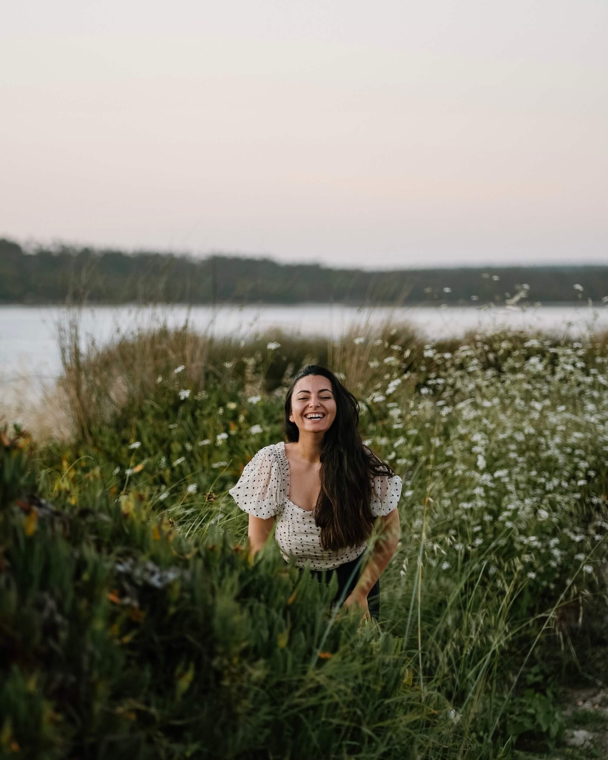 A woman with long dark hair smiling and laughing in a field of tall grass and white flowers near a body of water, with distant trees in the background.