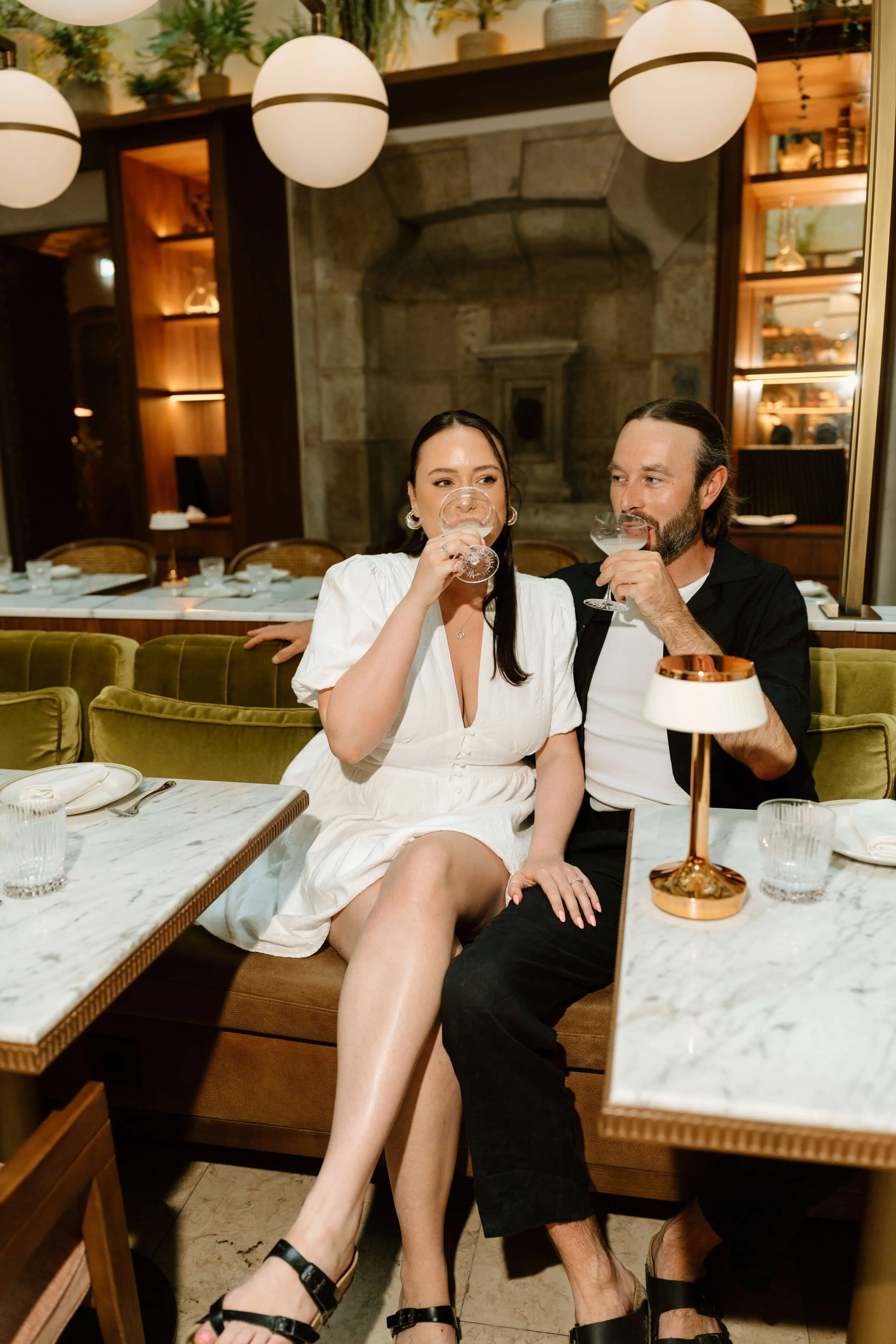 A couple sitting on a green velvet sofa at a restaurant, drinking cocktails and enjoying each other's company.