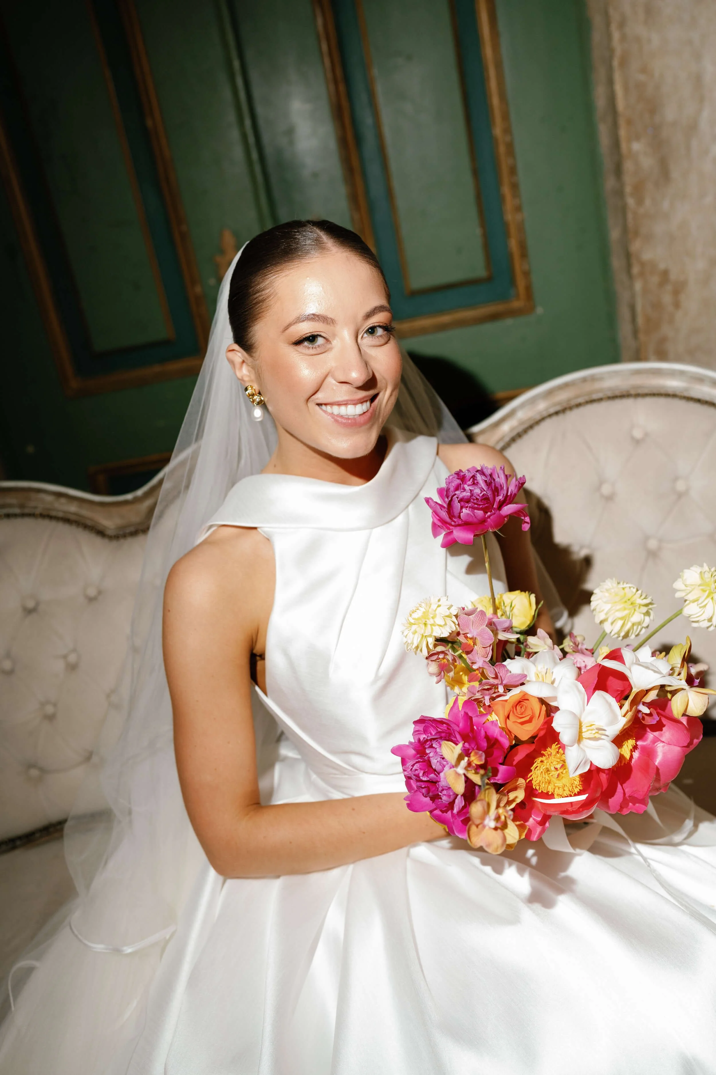 A smiling bride in a white wedding dress holding a bouquet of pink, white, yellow, and orange flowers, sitting on a vintage tufted cream-colored sofa.