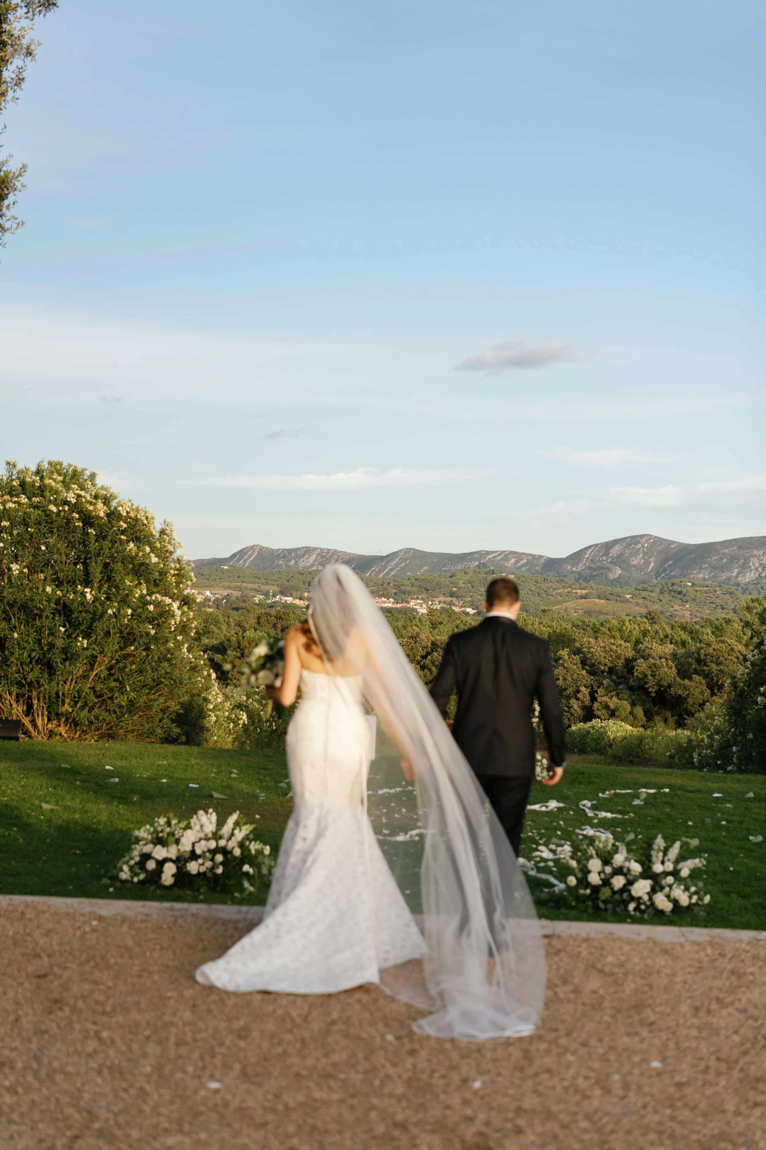 A bride and groom walking away together on a grassy hill with scenic mountains in the background, wedding flowers on the ground, during sunset.