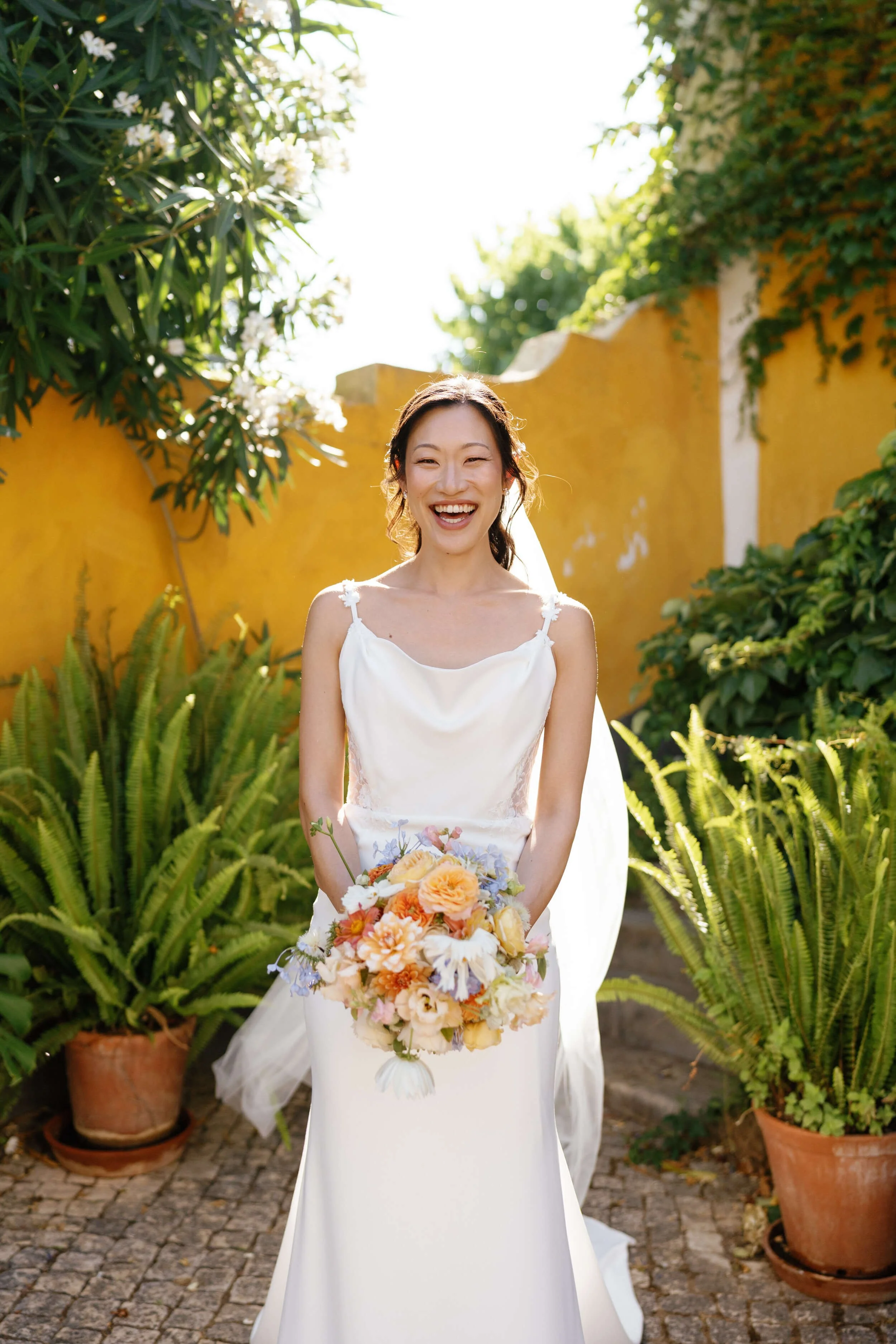 A smiling bride in a white wedding dress holding a colorful bouquet of flowers outdoors with yellow walls and potted green plants.