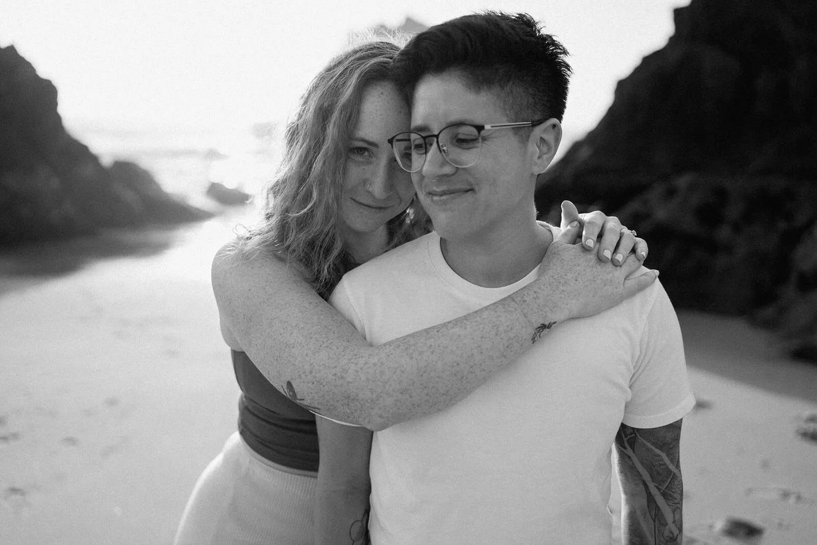 Same sex couple embrace on a beach with rocks in the background, smiling softly at each other in a black and white photo.