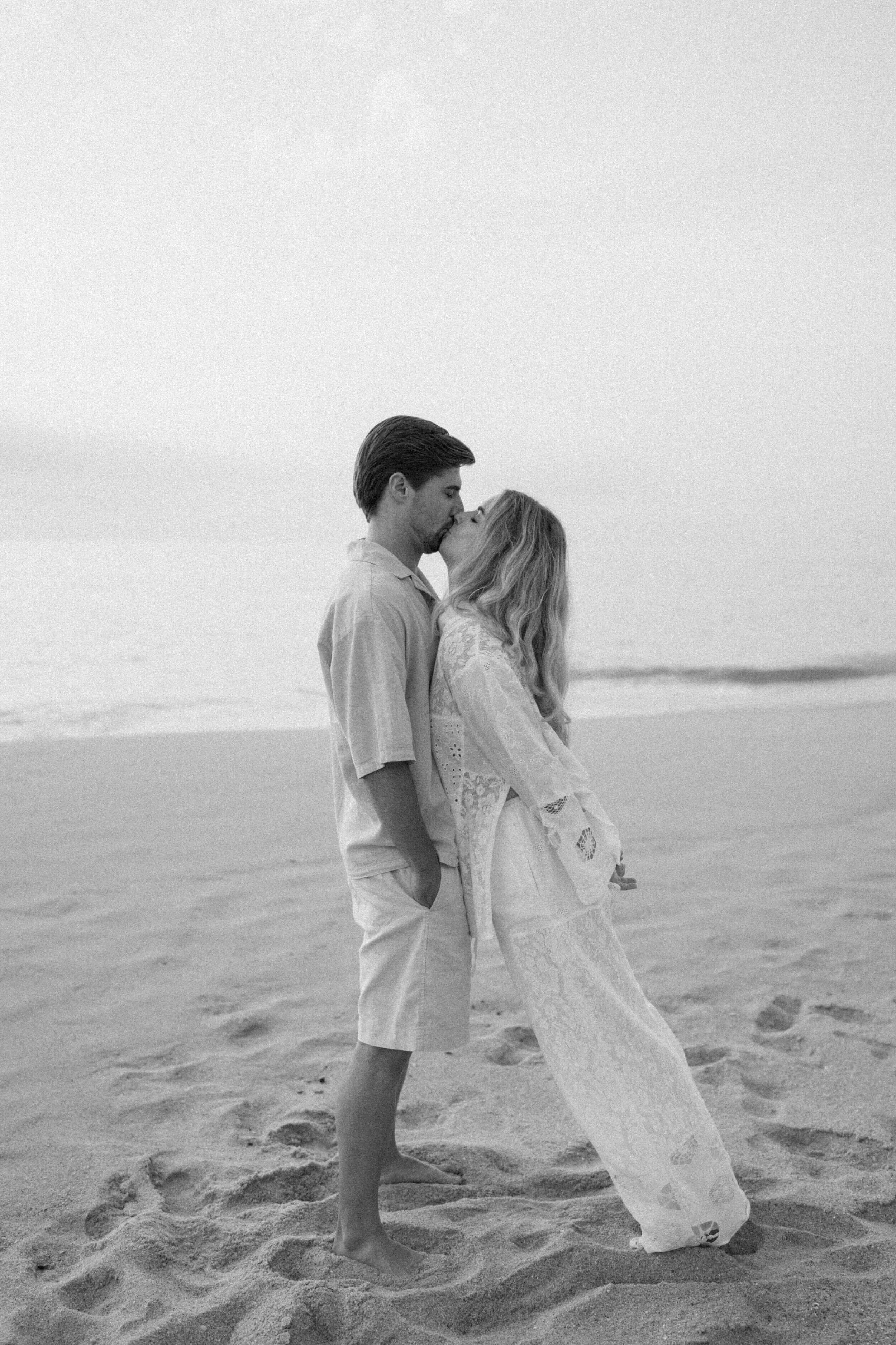A black and white photo of a couple kissing on the beach, standing close with the ocean in the background.