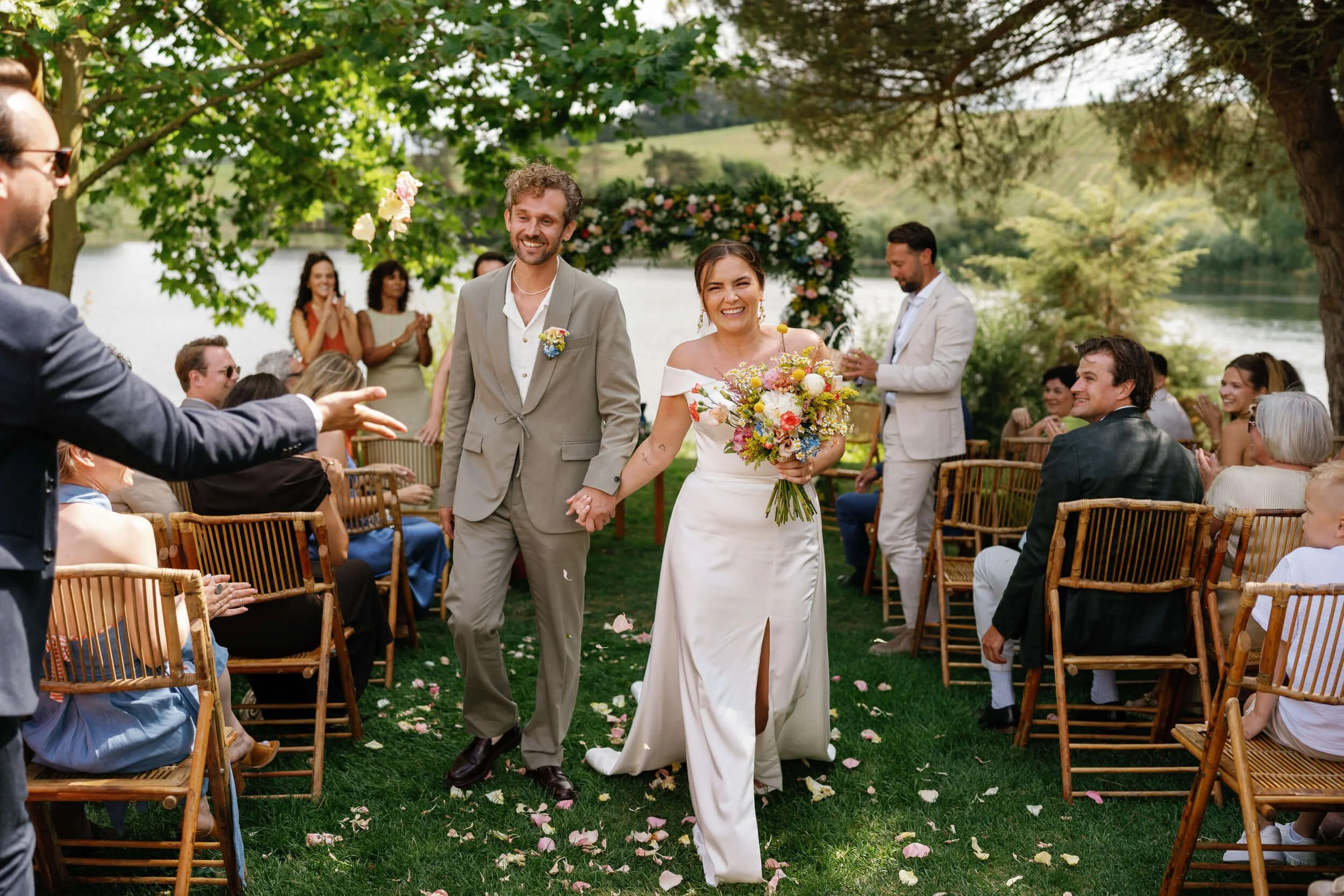 A newlywed couple walking hand-in-hand down an outdoor aisle, smiling, during their wedding ceremony by Quinta da Bichinha lake with guests seated on both sides, surrounded by trees and greenery.