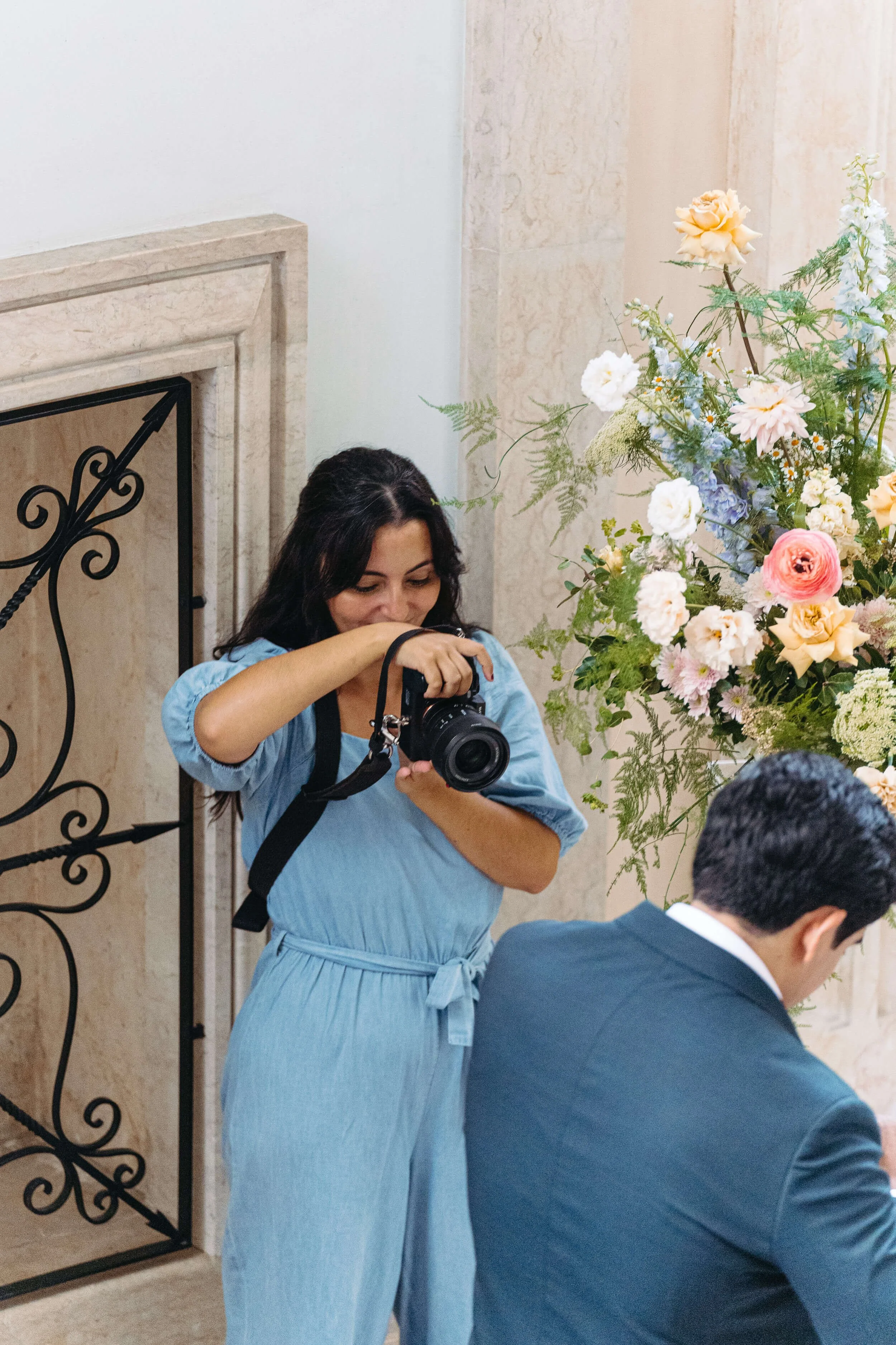 Woman with dark hair wearing a light blue dress holding a camera and looking down near a man with dark hair in a suit. A large floral arrangement of pink, white, and blue flowers is on the right side of the image.