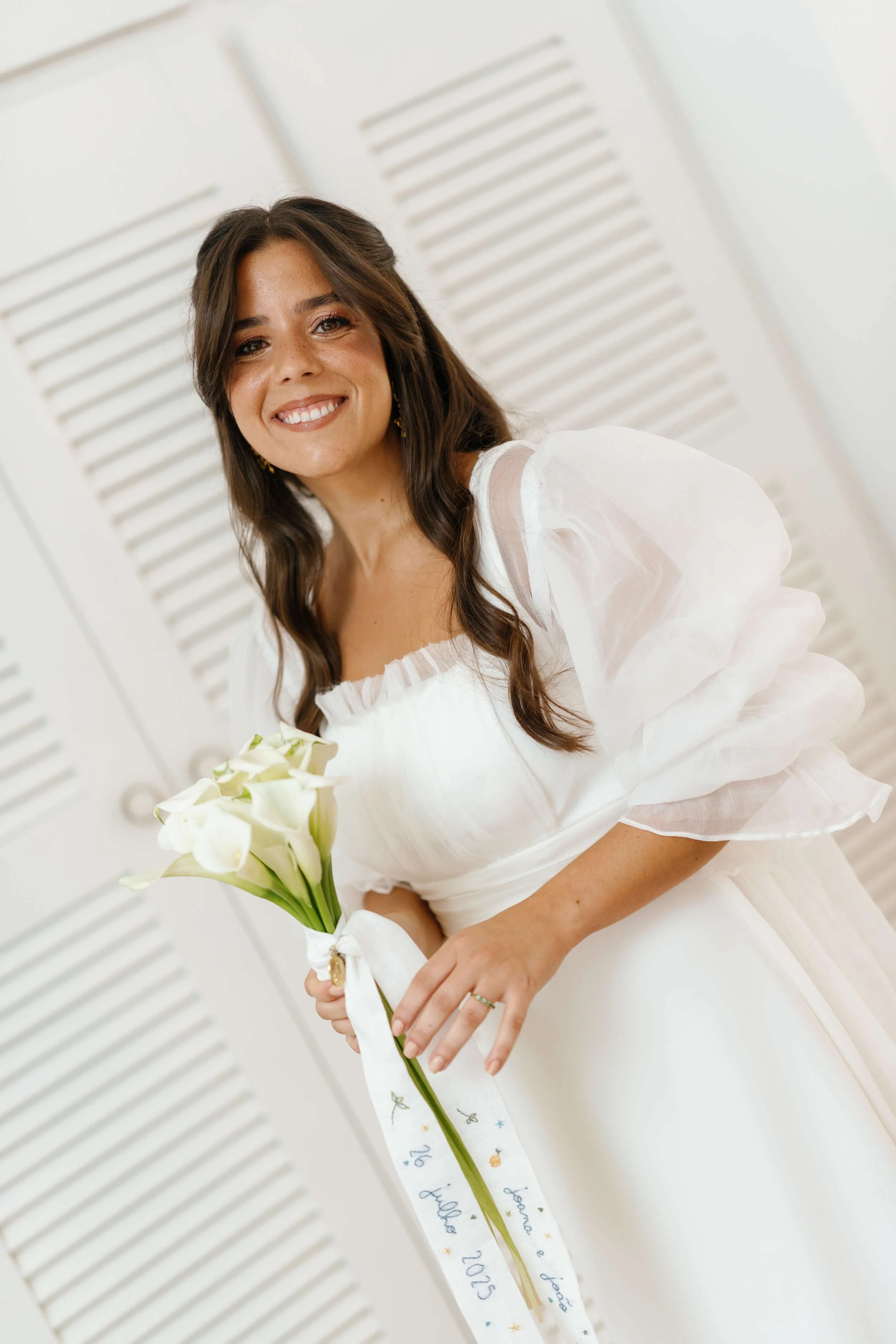 A smiling woman in a white dress holding a bouquet of white calla lilies with a ribbon that says 'le happy 2023'.