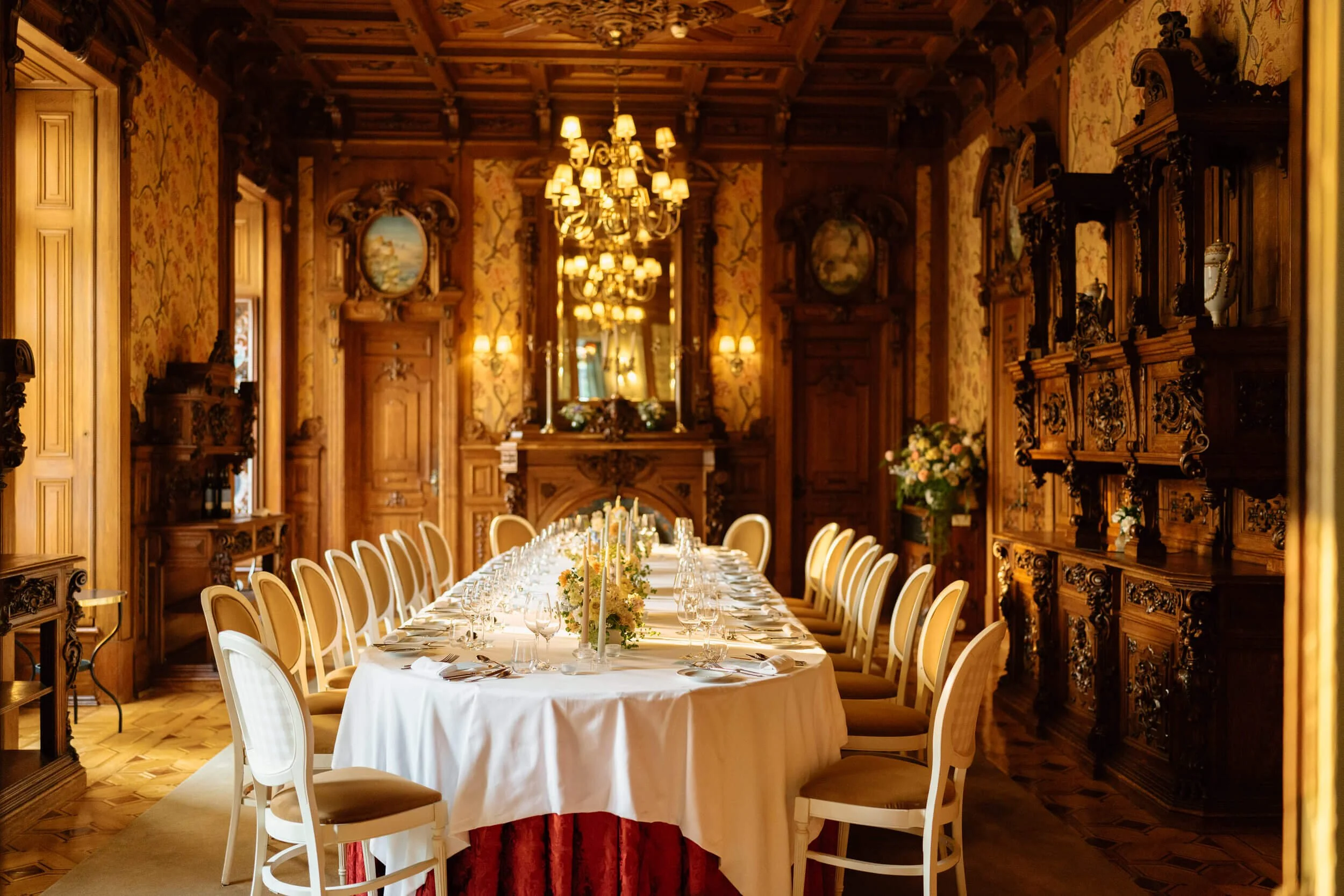 Elegant dining room with wood-paneled walls, chandelier, and a long table set for a formal dinner with floral centerpieces and candles.