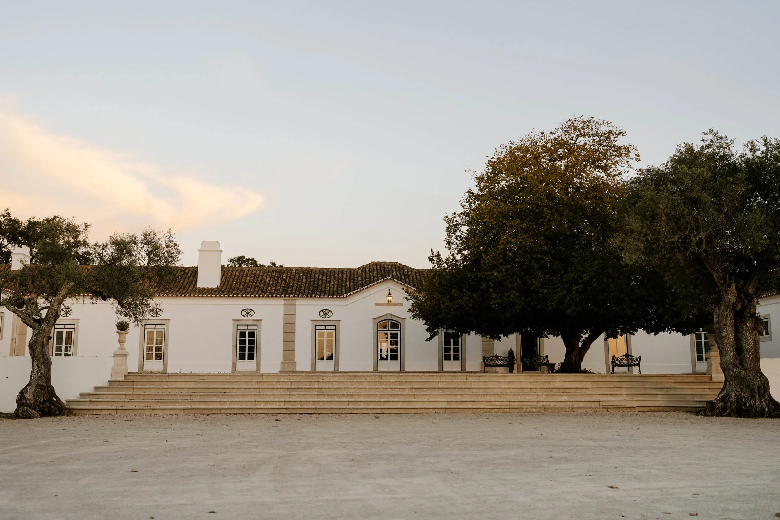 A white colonial-style building with steps leading up to the entrance, flanked by large trees and benches in front, under a partly cloudy sky during daylight.