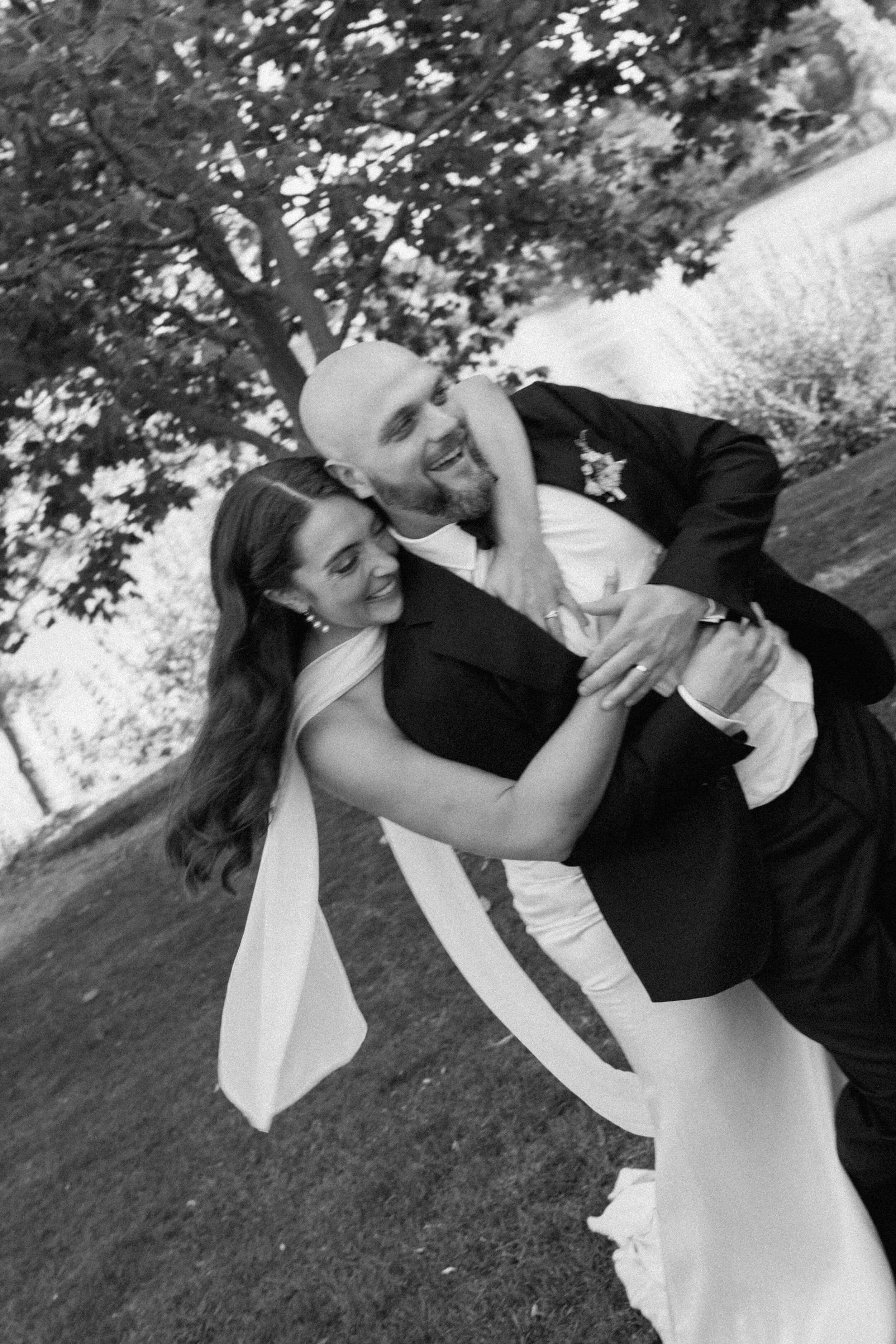 Black and white photo of a smiling couple in wedding attire, embracing outdoors near a tree.