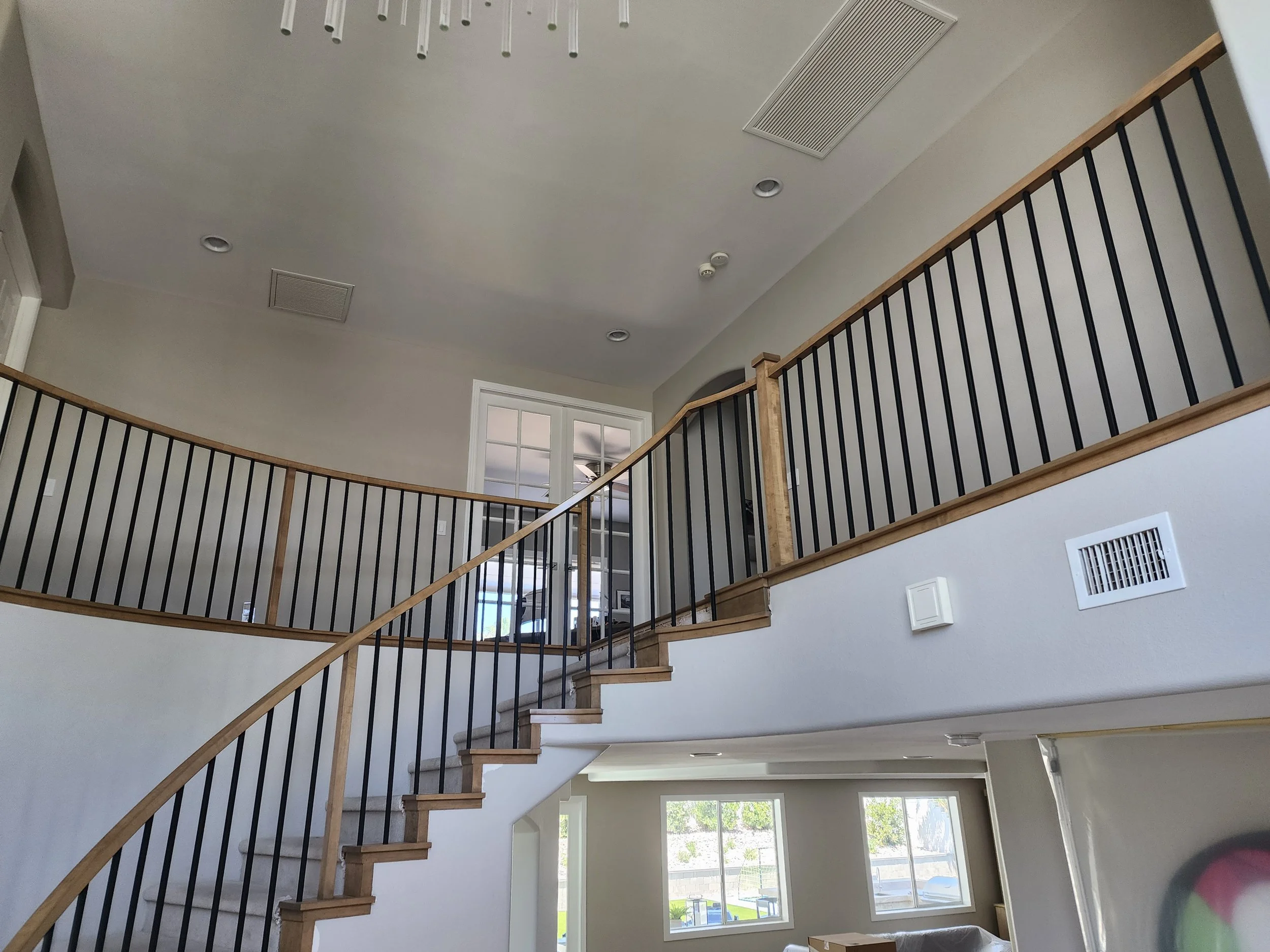Interior view of a spacious, modern home with a high ceiling, large windows, a staircase with wooden handrails and black metal balusters, and ceiling vents visible.
