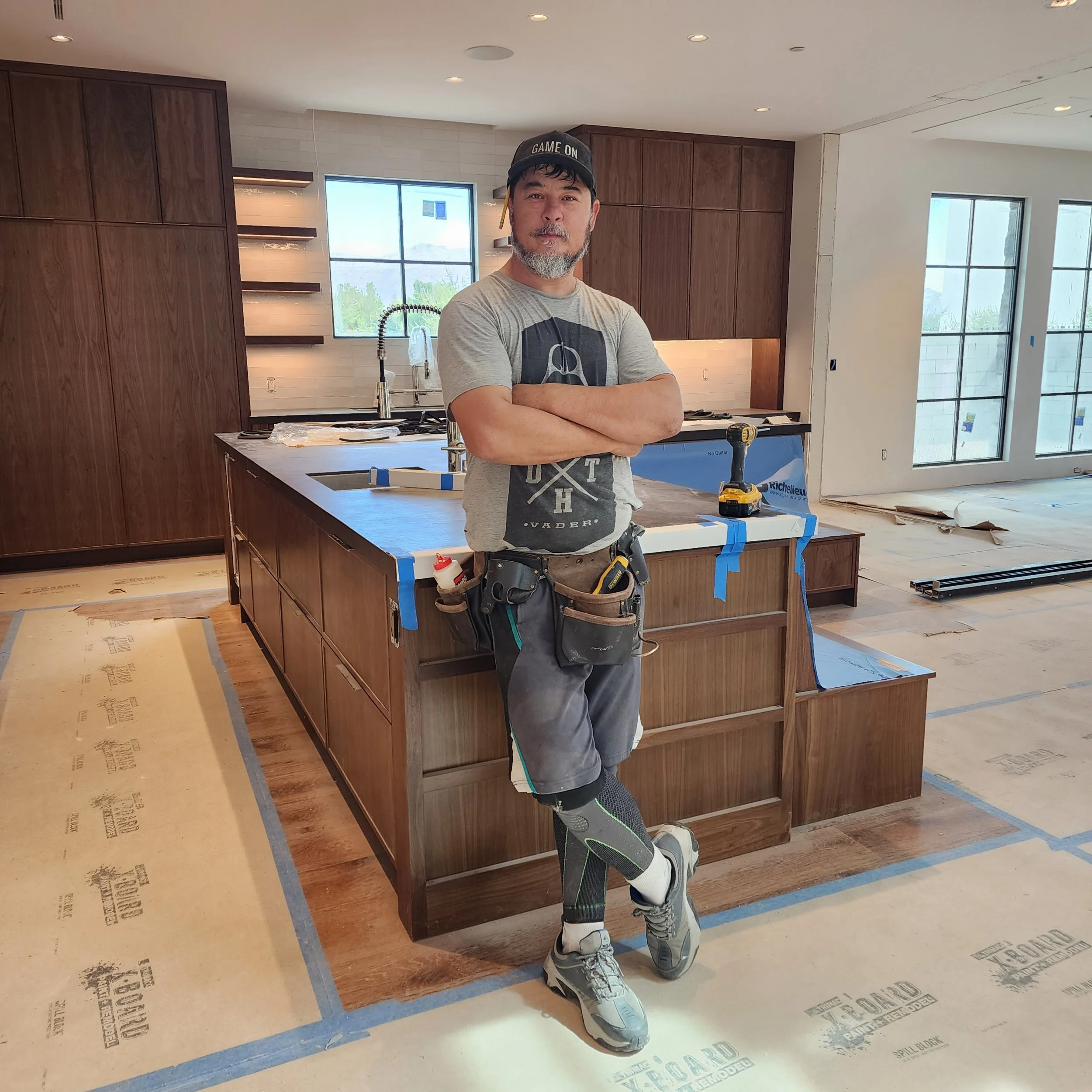 A master carpenter in a kitchen under construction. The kitchen has dark wooden cabinets, a large window, and an island with construction tools around.