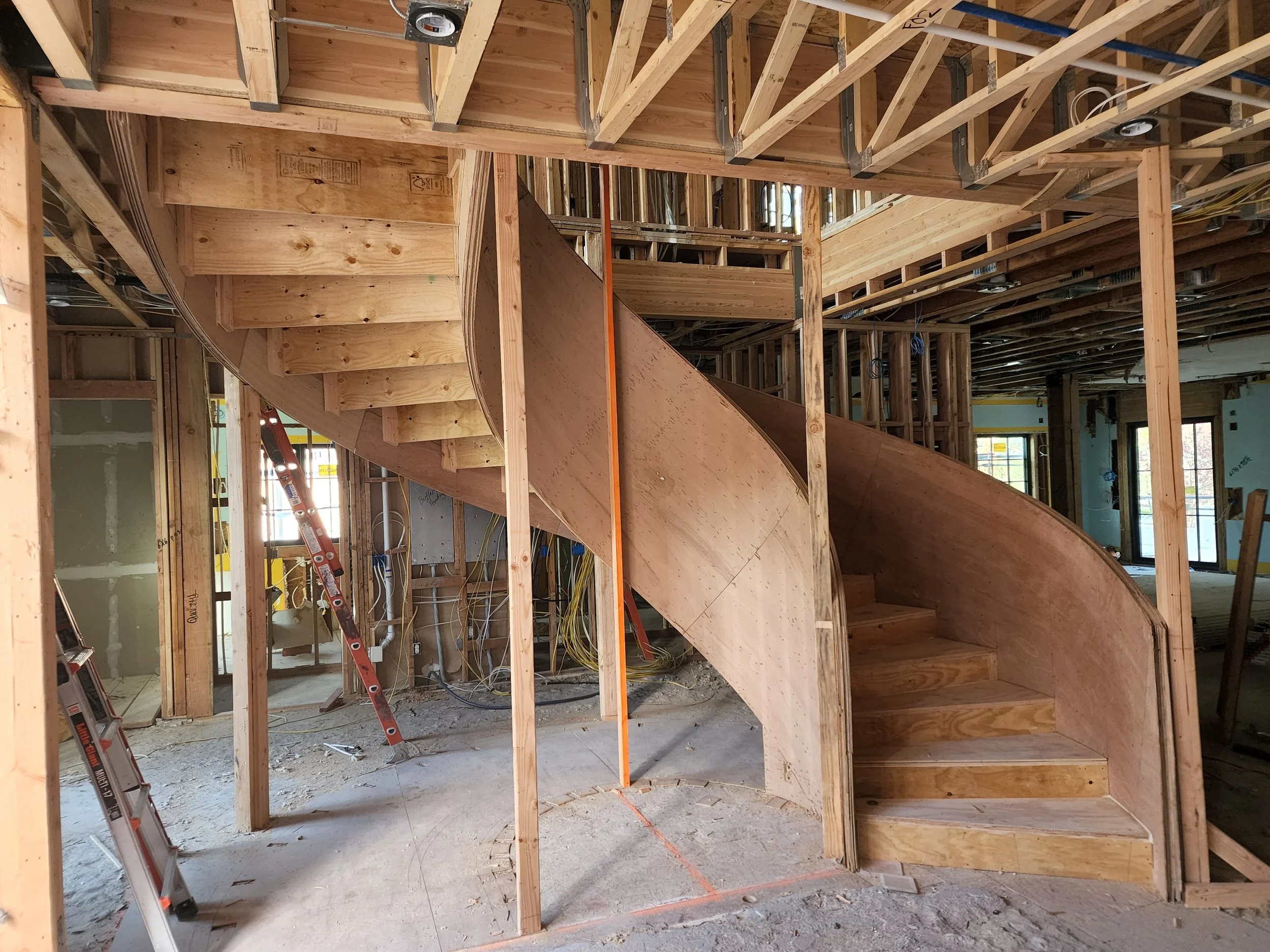 Inside a building under construction with exposed wooden framing, stairs, and electrical wiring. Ladders and construction tools are visible.