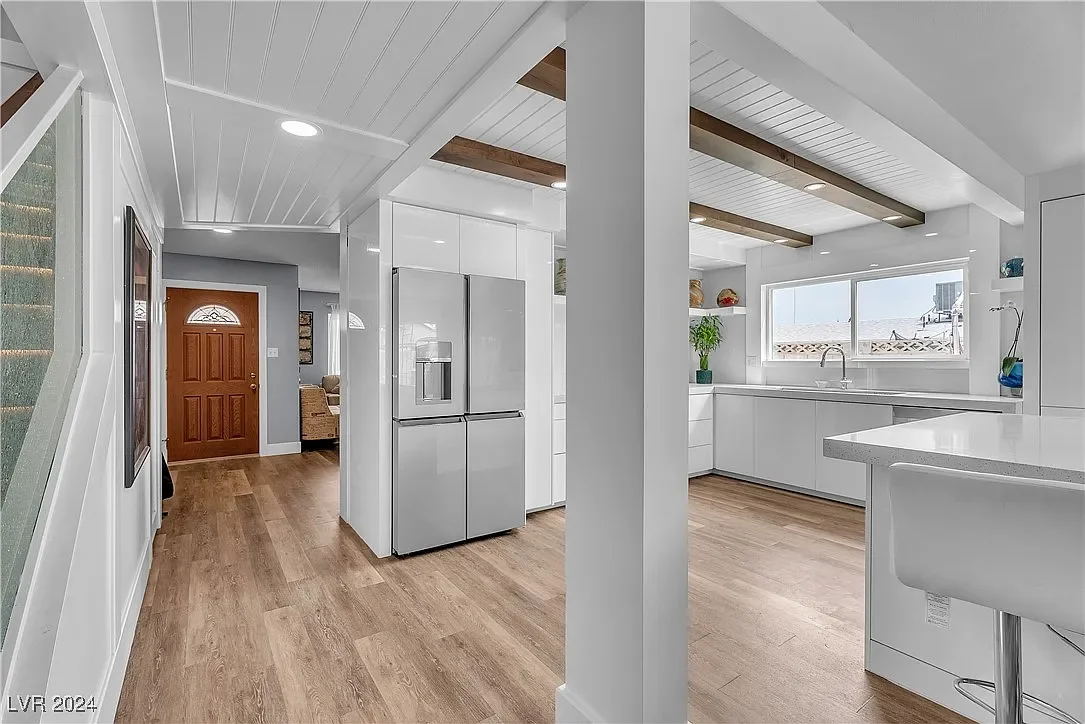 Bright, modern kitchen with white cabinets, stainless steel refrigerator, and wood flooring, featuring a large window over the sink and decorative open shelves.