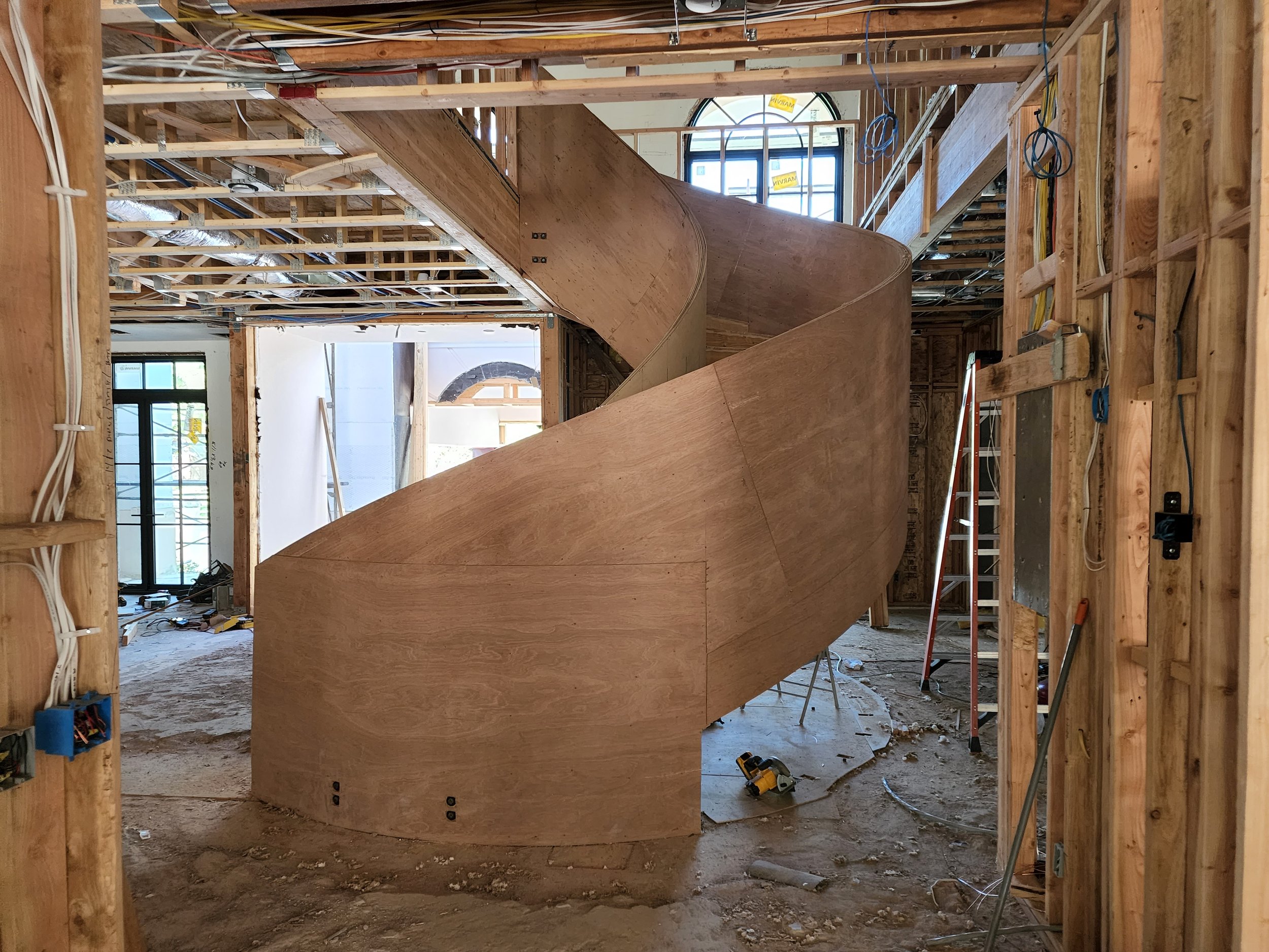 Interior view of a building under construction with exposed wooden framing, stairs, and electrical wires, and large windows letting in natural light.