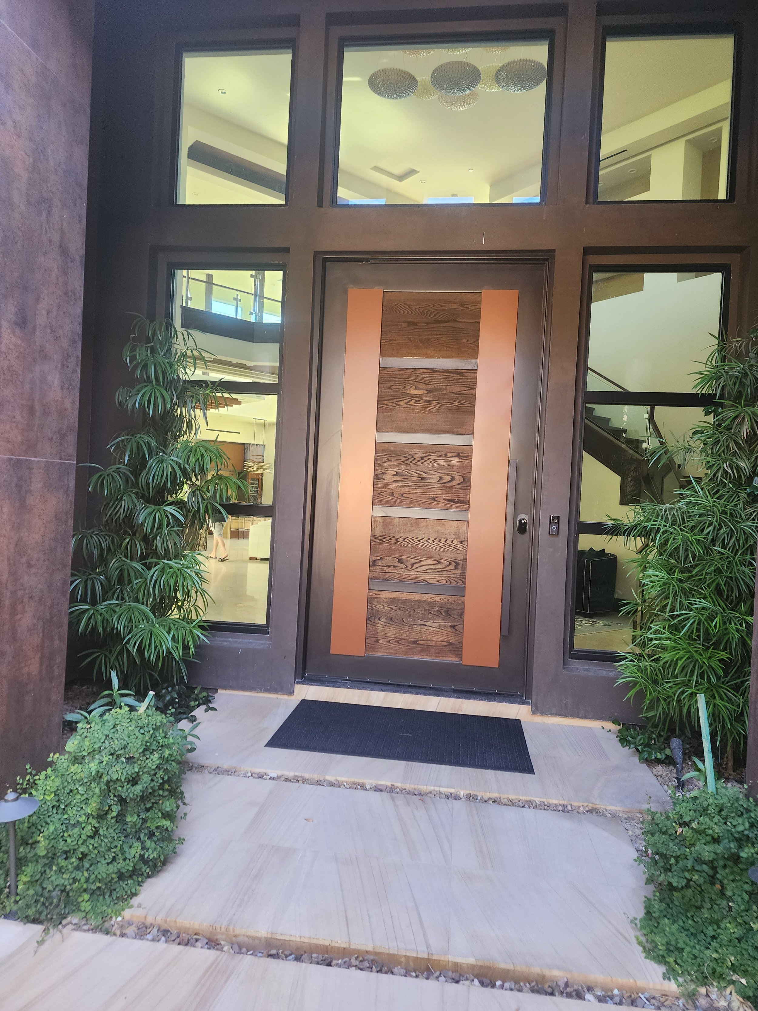 Modern front door of a building with large glass windows on either side, a wooden panel design, and a black doormat. Green plants surround the entrance with a manicured garden bed and concrete walkway.