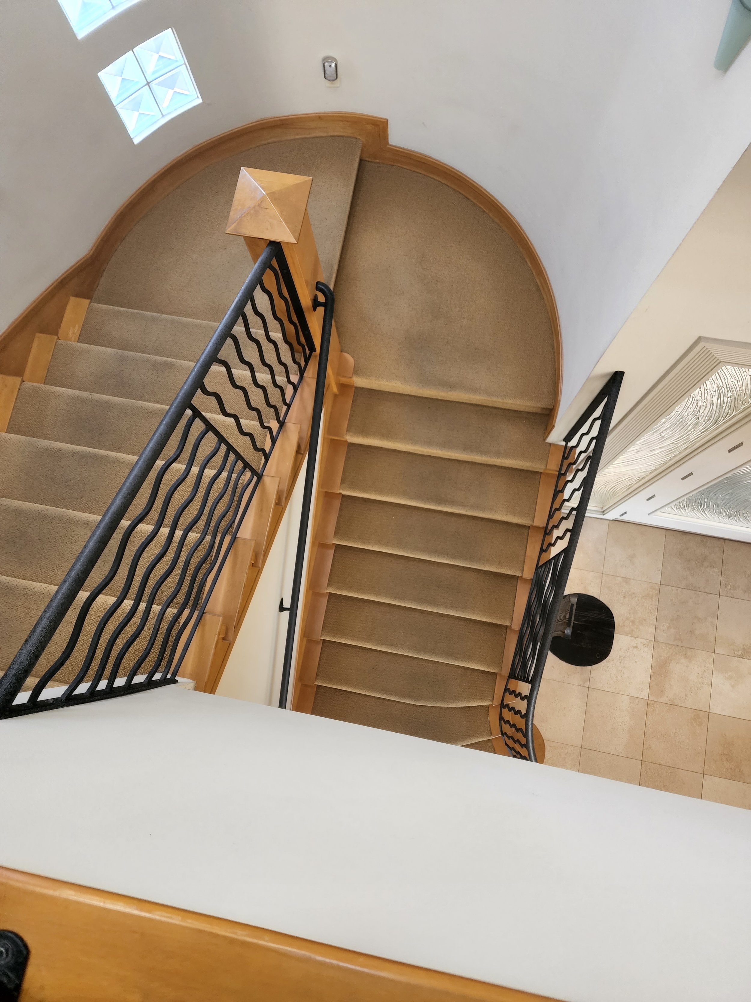 A spiral staircase with beige carpet steps, black metal railing, wooden handrail, and a small glass block window in a white wall, viewed from above.