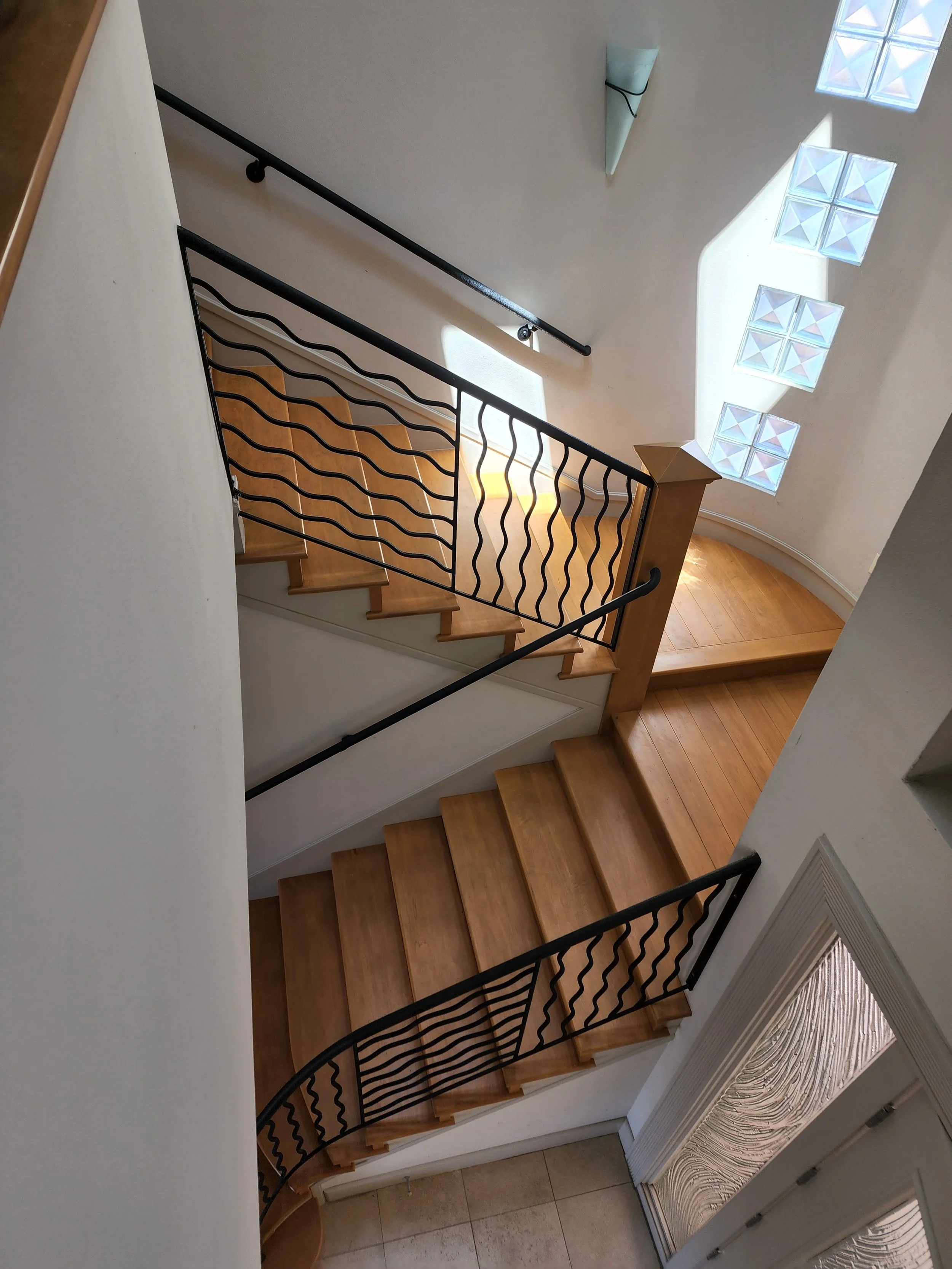 Interior view of a staircase with wooden steps, black metal railings, white walls, a wall-mounted light fixture, and a glass window allowing sunlight to shine through.