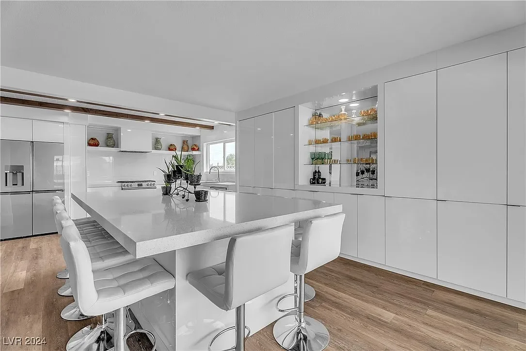 Modern white kitchen with an island, barstools, and built-in glass cabinets displaying glassware.
