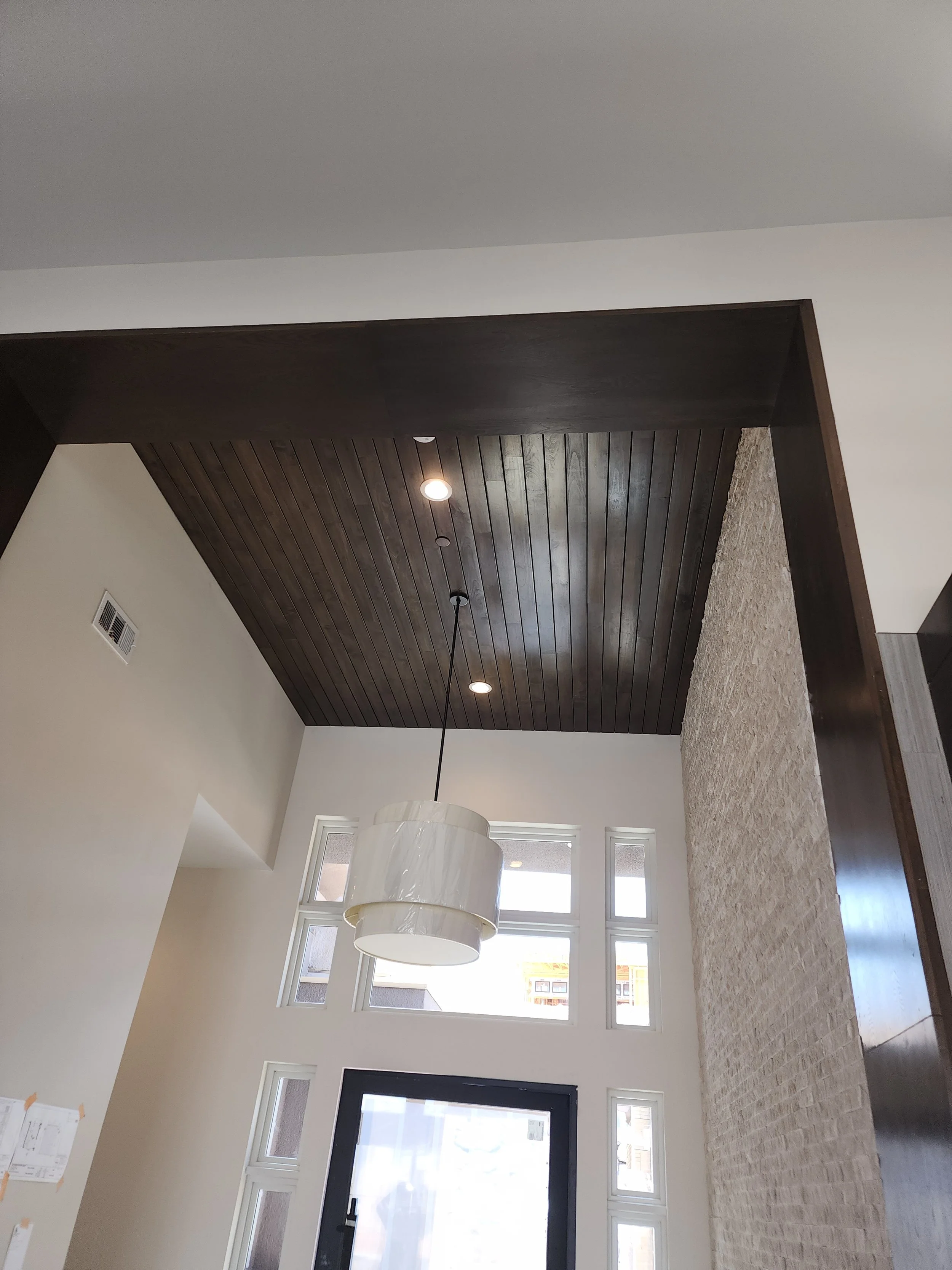 Interior view of a modern living space with a dark wooden ceiling, beige carpeted floor, white walls, and large windows allowing natural light.