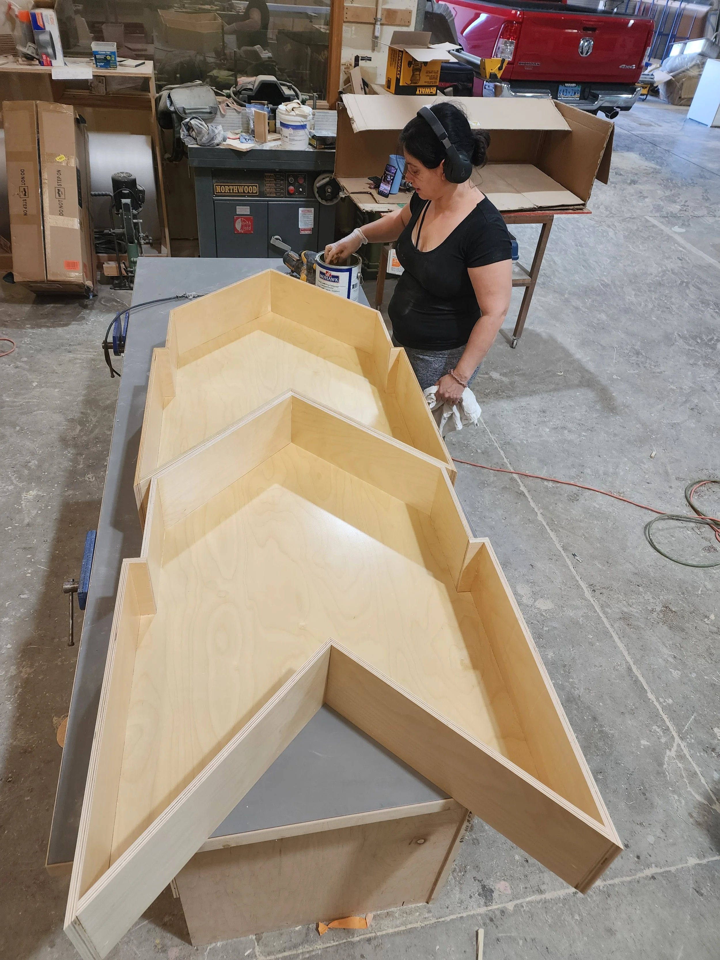 A woman is working in a workshop, assembling a large wooden star-shaped frame. She is wearing headphones, gloves, and a black top. The workshop has various tools, boxes, and a red truck in the background.