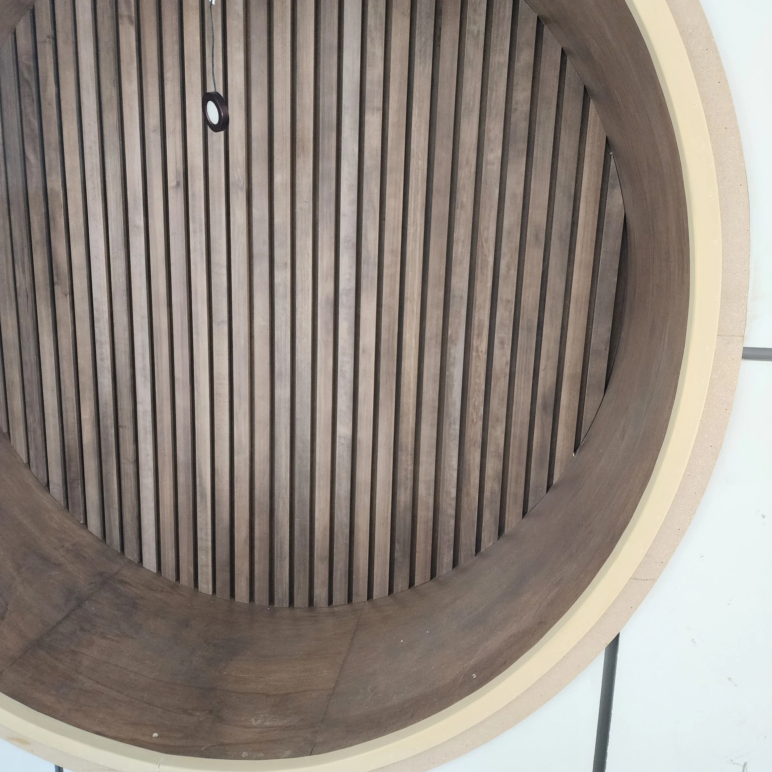 Ceiling with wooden slats and a round light fixture, viewed from below, with a curved wooden border and partial view of a beige wall.