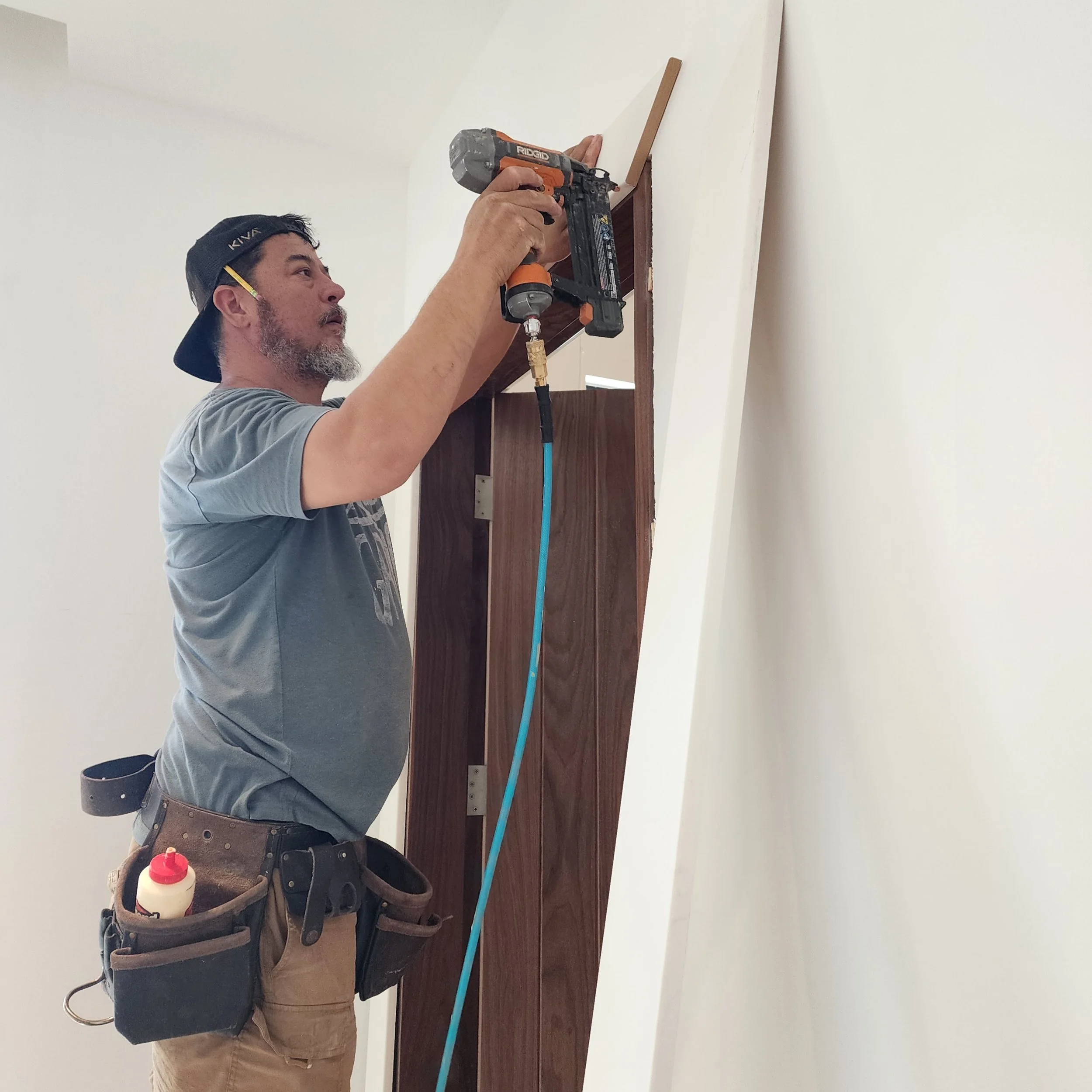 A man wearing a black cap backwards, gray t-shirt, and tool belt is using a cordless nail gun to attach wood trim to a wall.