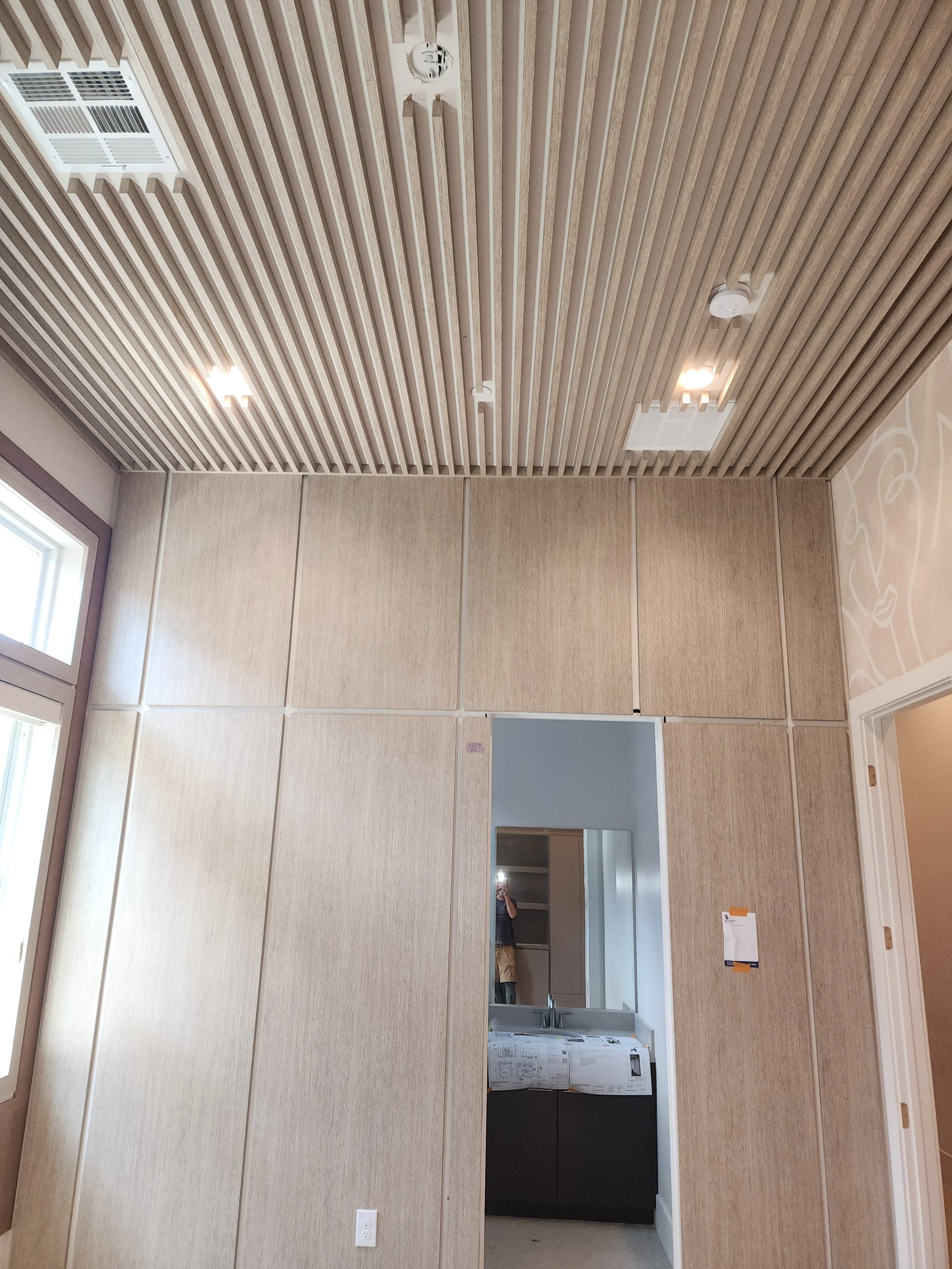 Photo of an interior wall with wood paneling, ceiling with wood slats, ceiling lights, an air vent, and a mirror reflecting a person taking the photo.