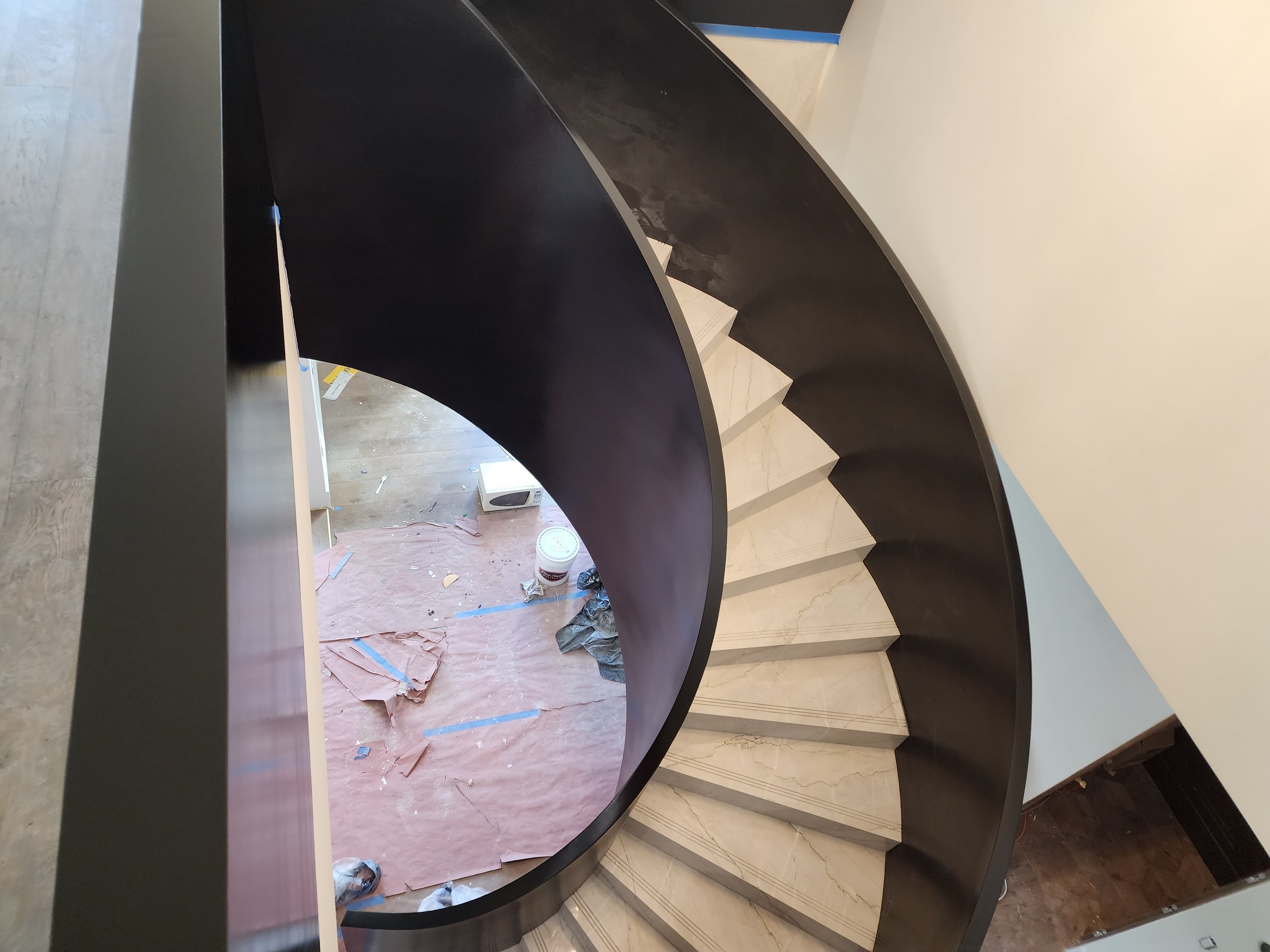 A black curved staircase with white marble steps and beige marble flooring, with construction materials and tools visible at the bottom of the stairs.