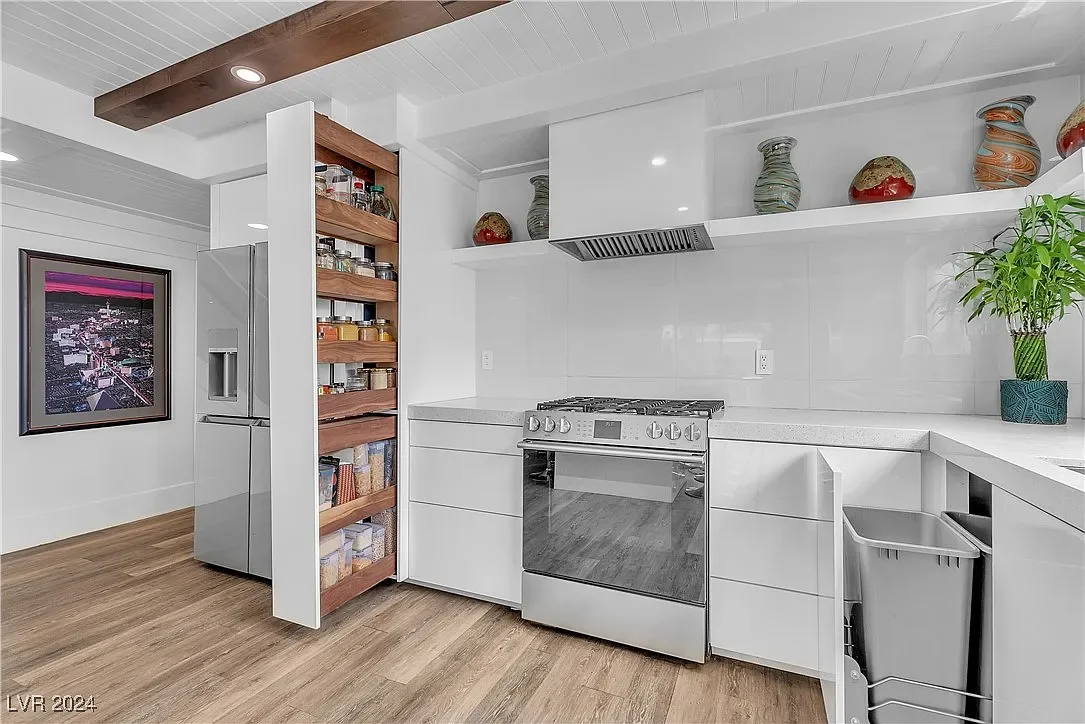 Modern kitchen with white cabinets, wooden shelves with spices, stainless steel oven, and refrigerator, decorated with vases and a potted plant, with a wooden ceiling beam and hardwood flooring.